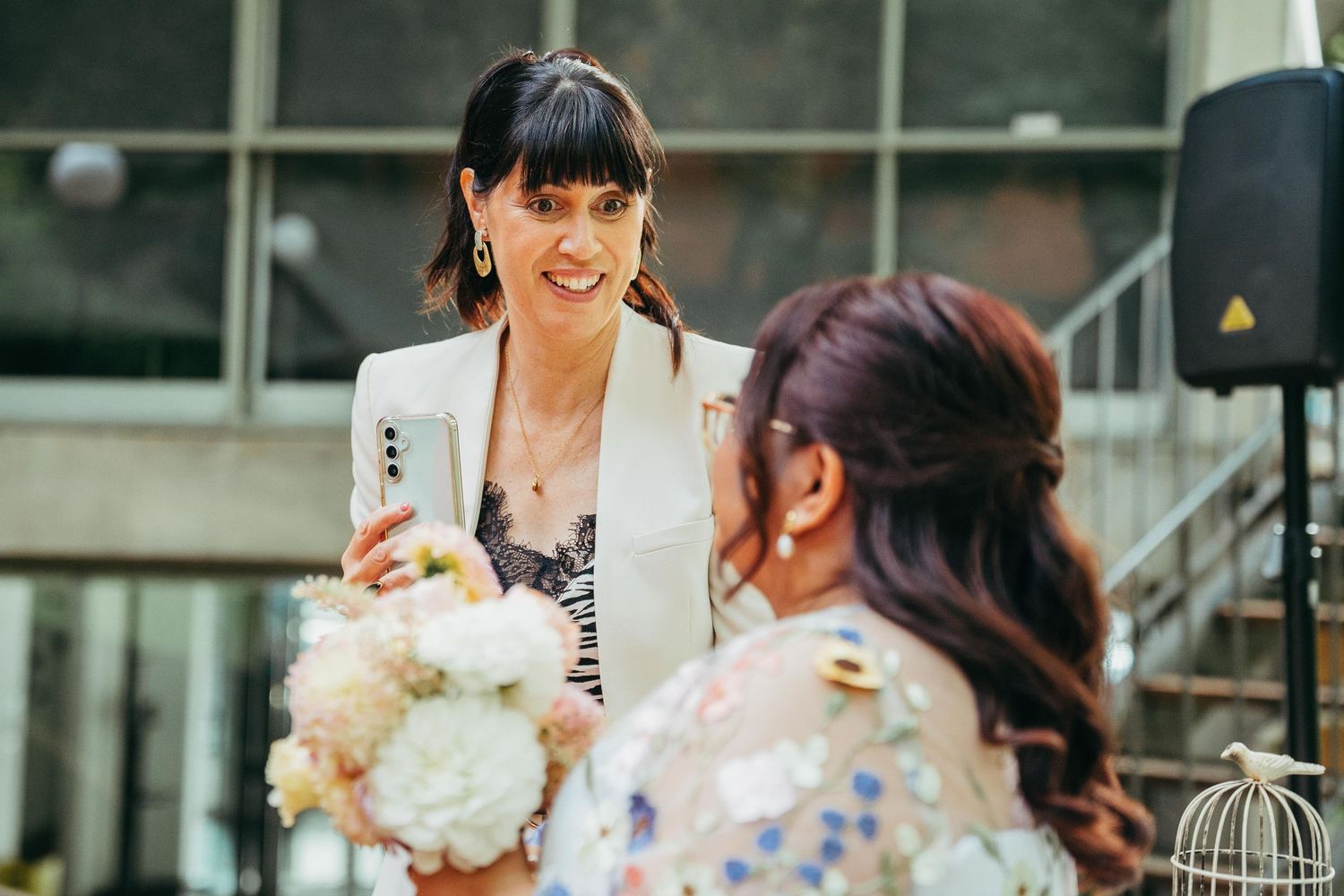 Woman in white blazer smiling at bride holding white bouquet at wedding.