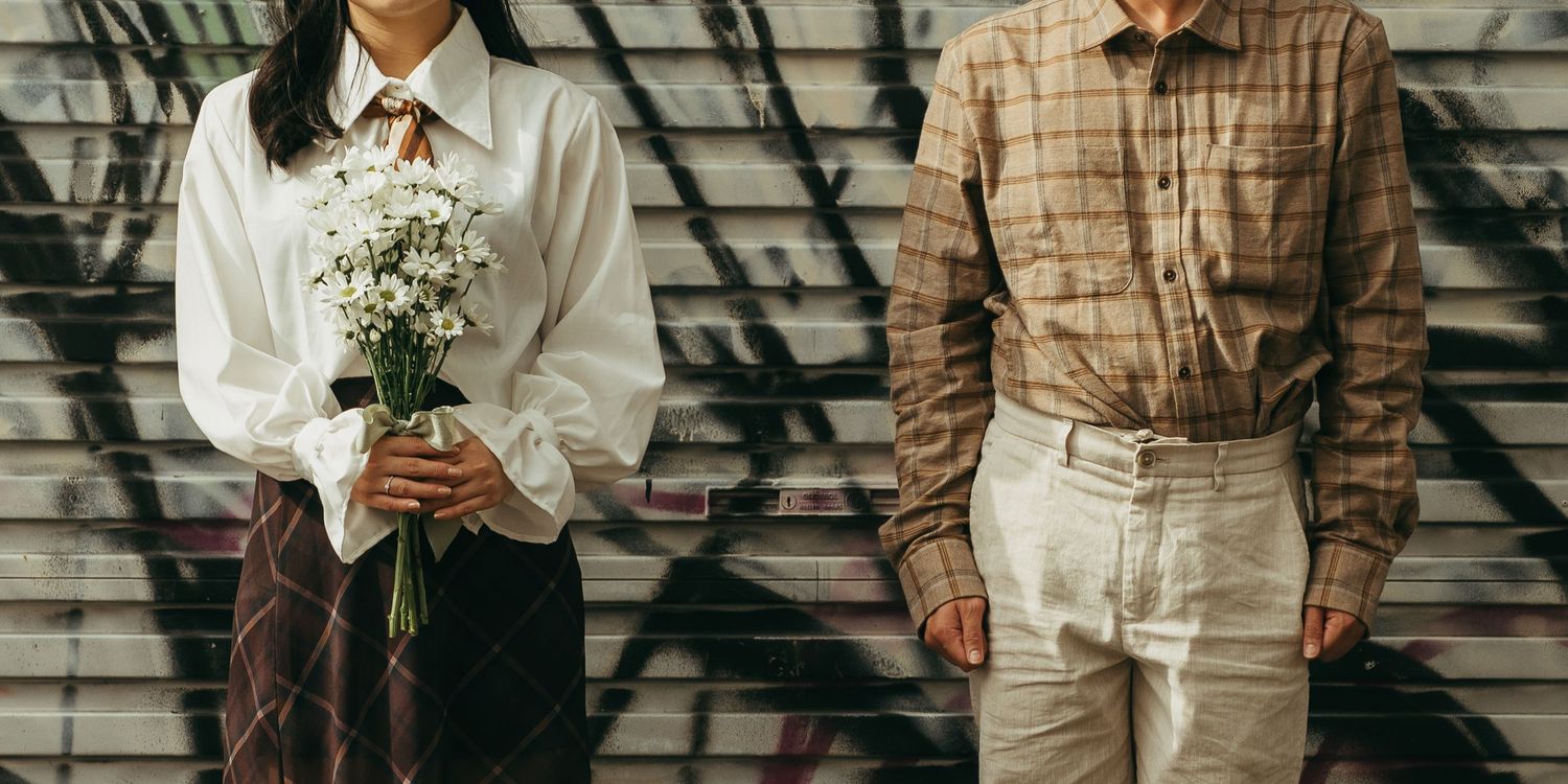 A couple stands before a graffiti wall, woman holding white flowers in skirt and blouse, man in plaid shirt and beige pants.