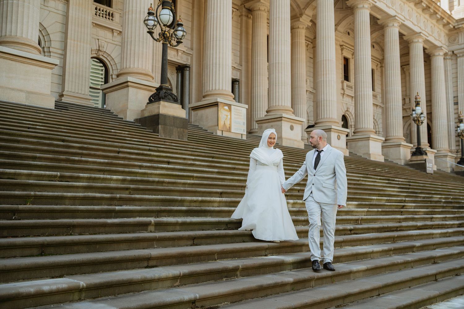 Muslim bride and groom in white wedding attire holding hands on grand stone steps of a classical building.