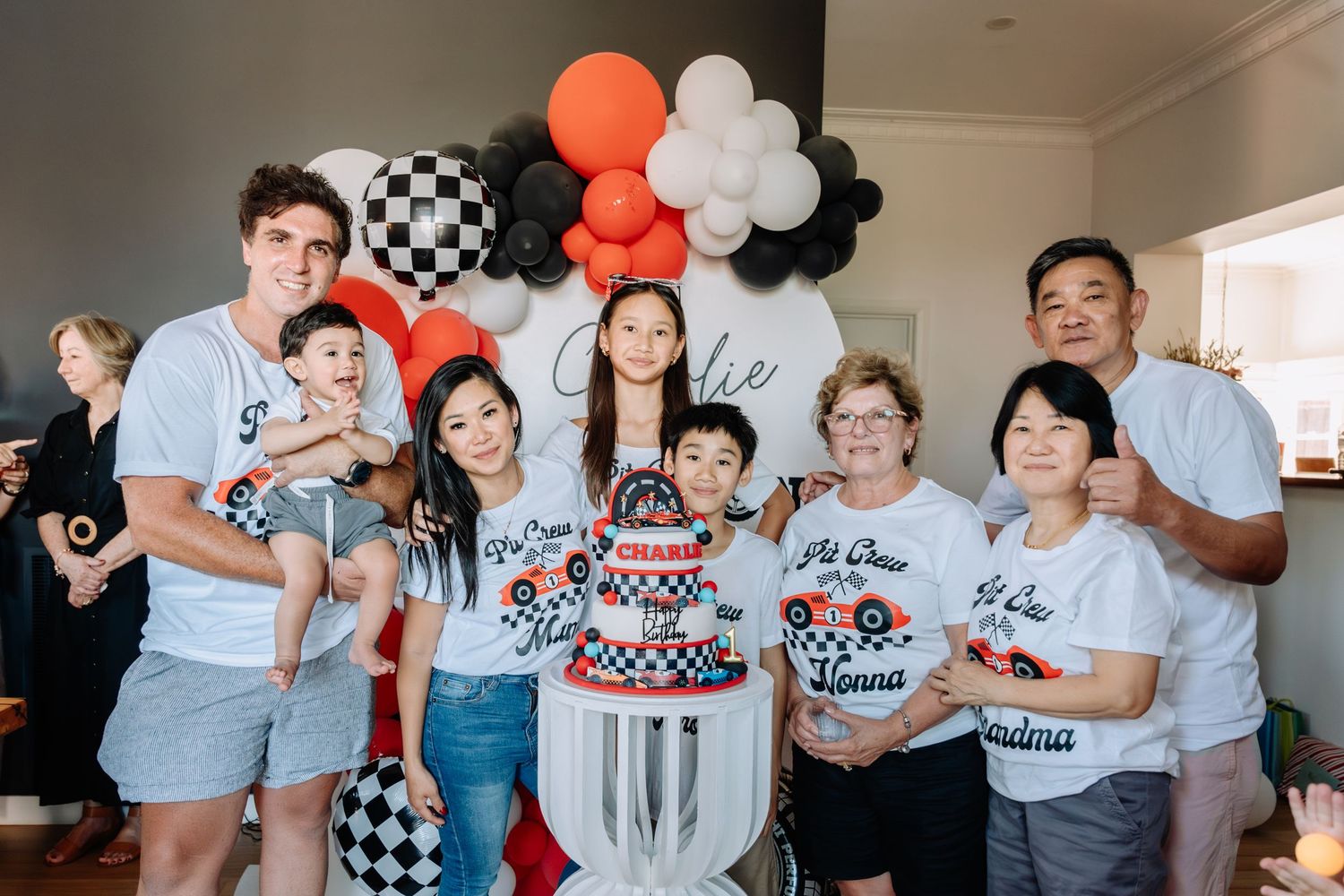 A group wearing matching racing-themed t-shirts celebrates at a party with black, orange and white balloon decorations.