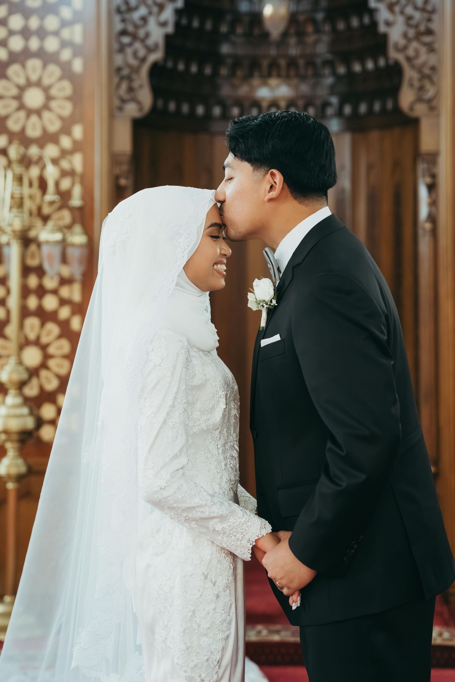 A groom in a black suit kisses the forehead of his bride in a white hijab inside an ornate mosque.