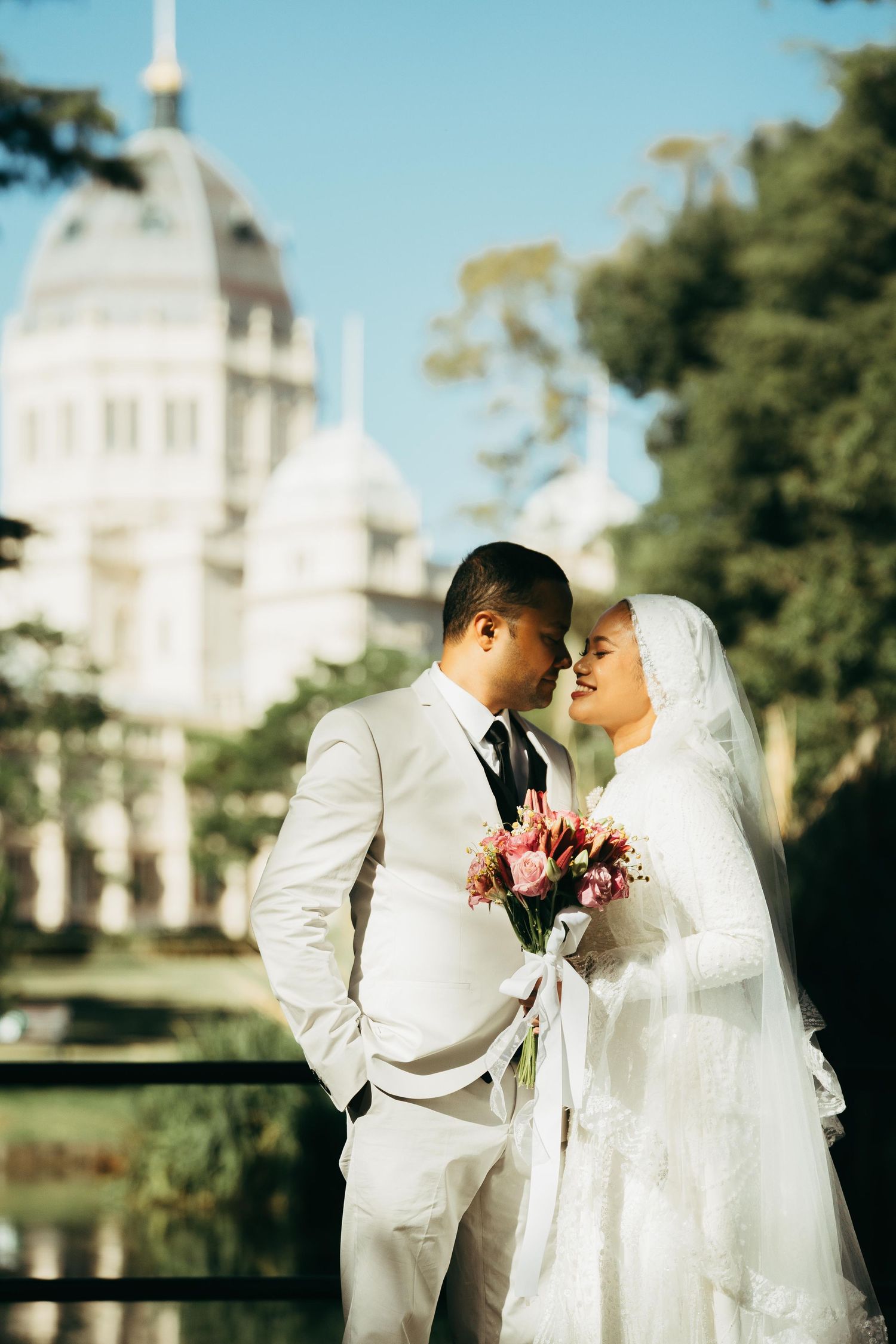 Muslim wedding couple in white attire sharing romantic moment with bouquet, historic building backdrop.