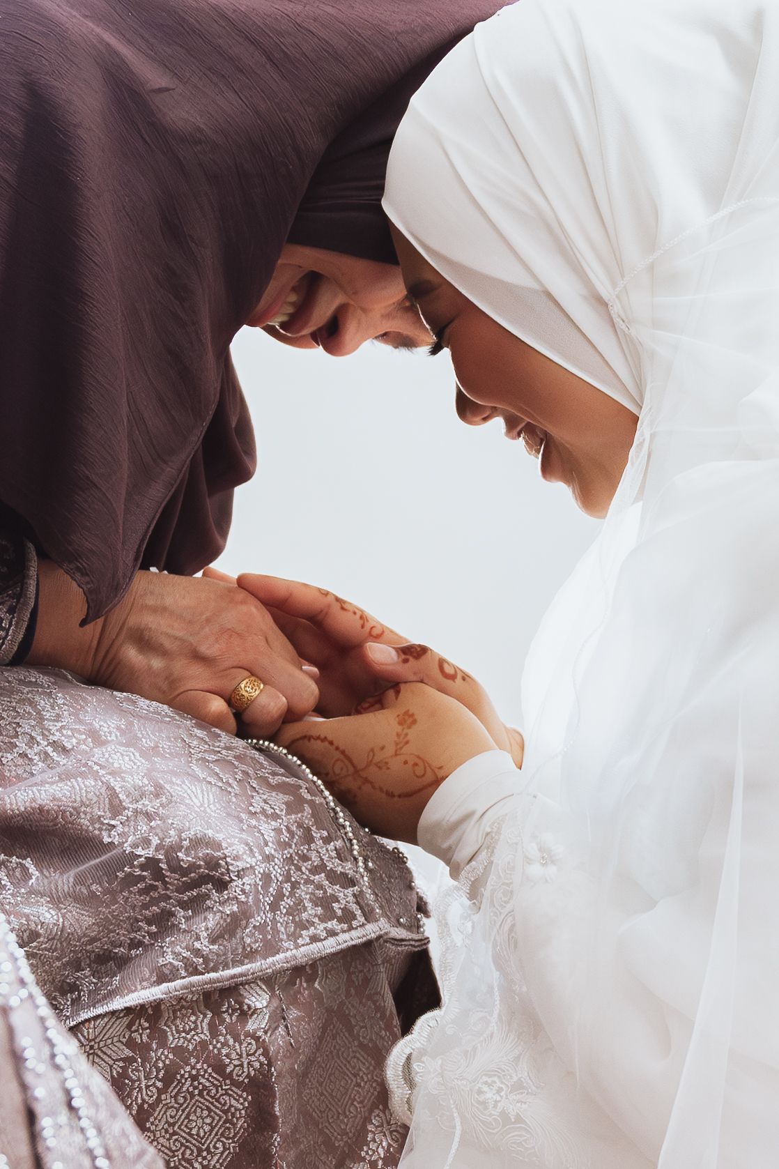 Two Muslim women in hijabs share a tender moment, touching foreheads while holding hands adorned with henna.