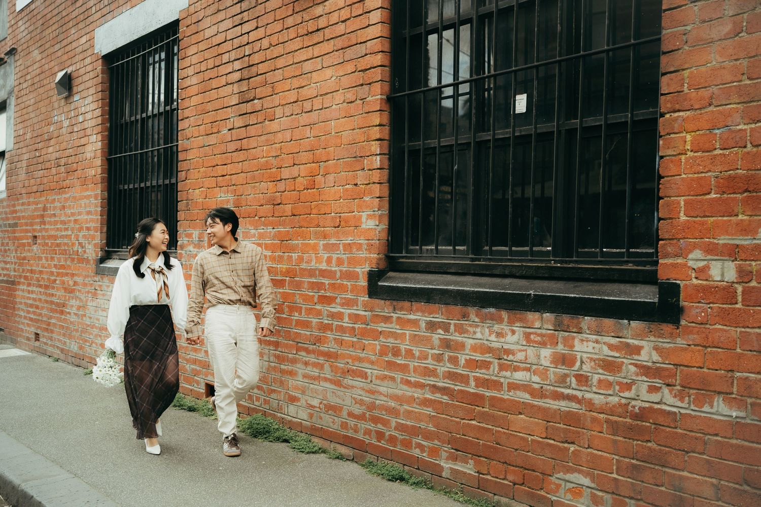 Couple walking hand in hand along a brick wall street, smiling at each other in casual outfits.