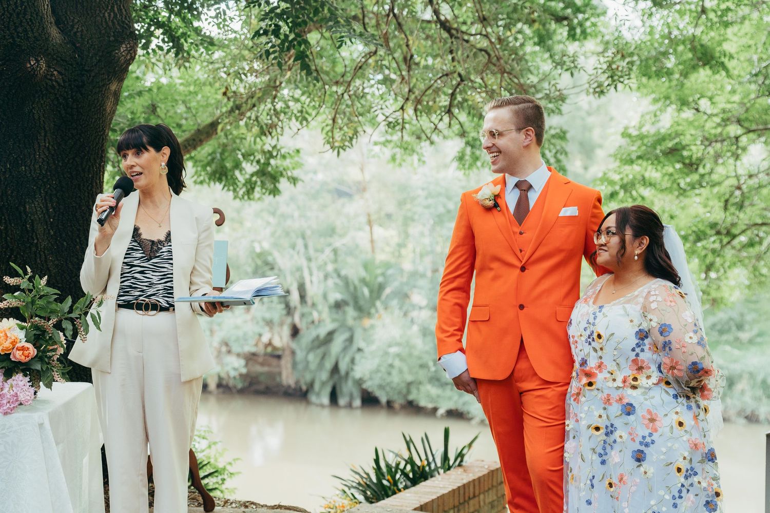 Groom in bold orange suit and bride in floral dress stand with officiant during outdoor wedding ceremony.