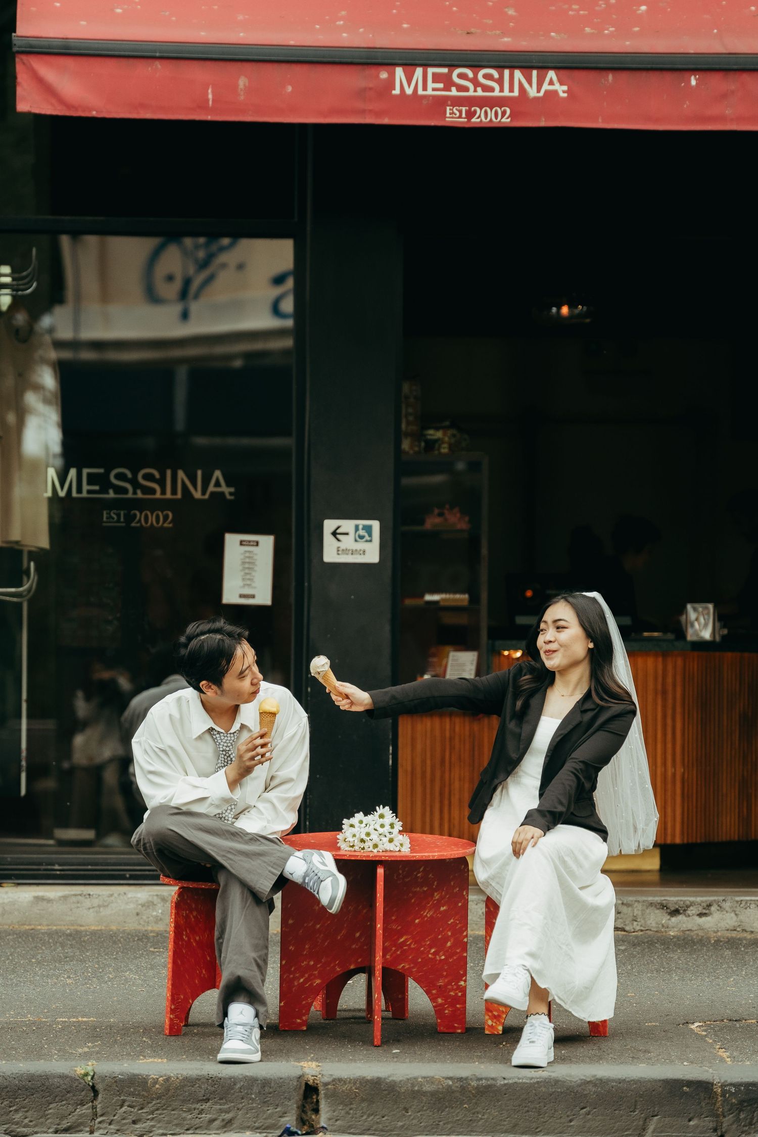 Couple sharing ice cream at a red table outside Messina gelato shop, woman in bridal attire offering a cone to her partner.
