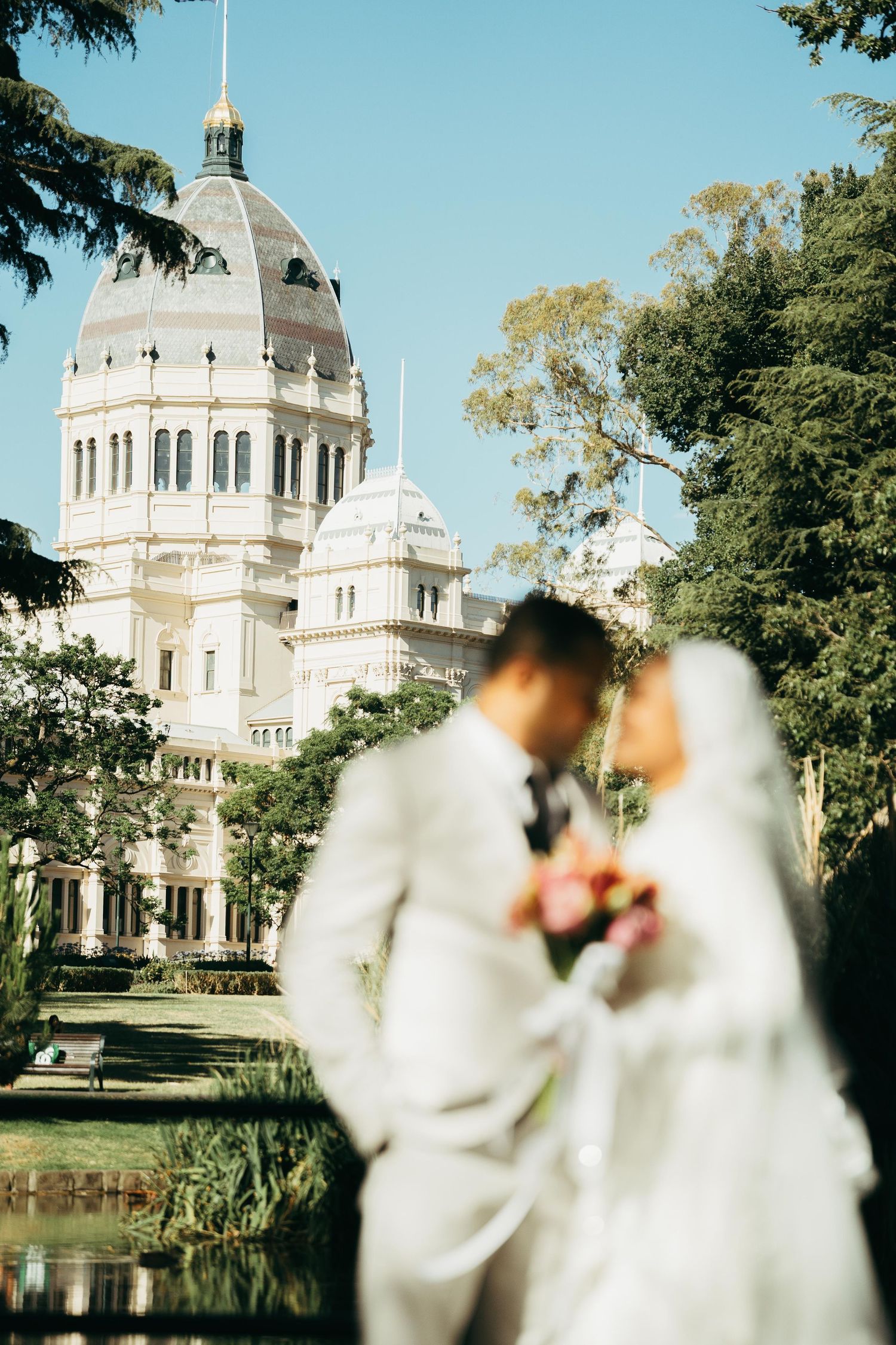 Bride and groom pose in front of a grand domed building surrounded by lush trees on a sunny wedding day.