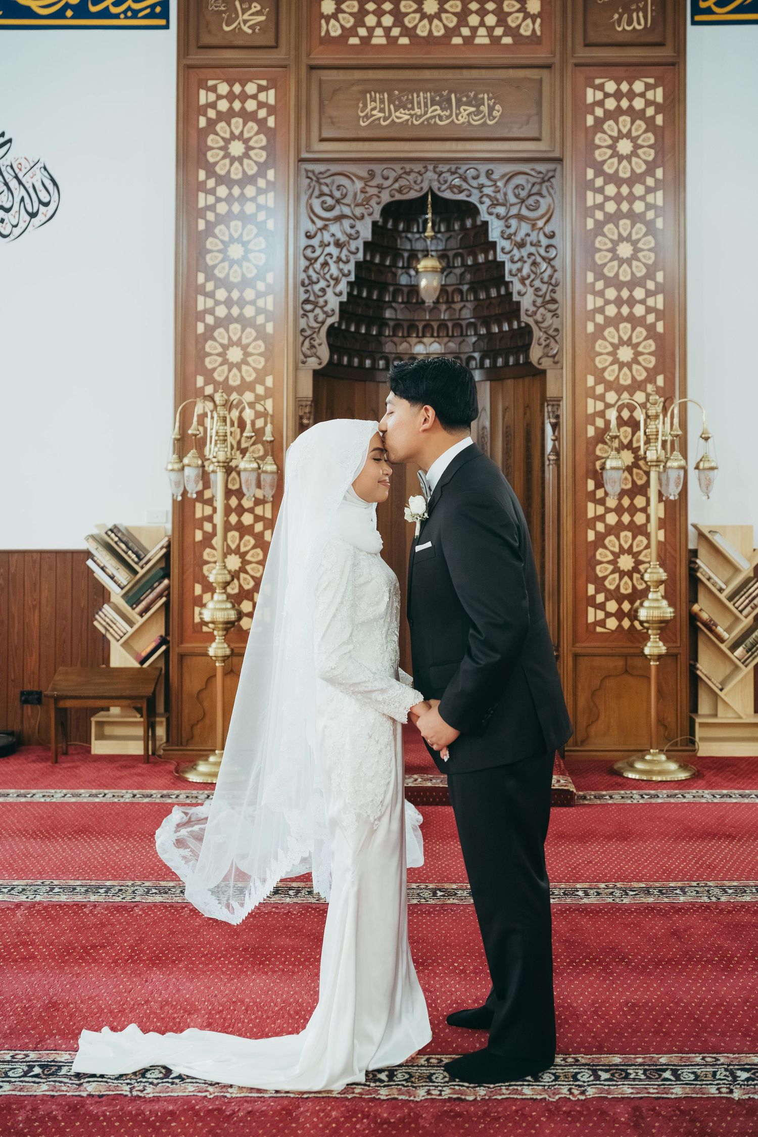 Muslim bride and groom sharing intimate forehead kiss before ornate mosque mihrab.
