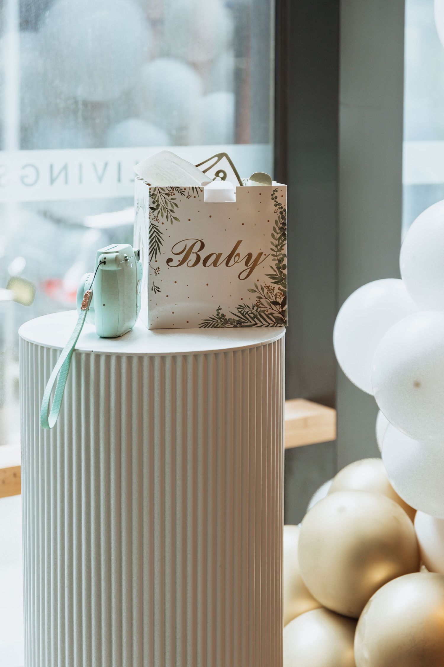 An elegant baby gift bag sits on a modern cylindrical pedestal with white balloons in the background.