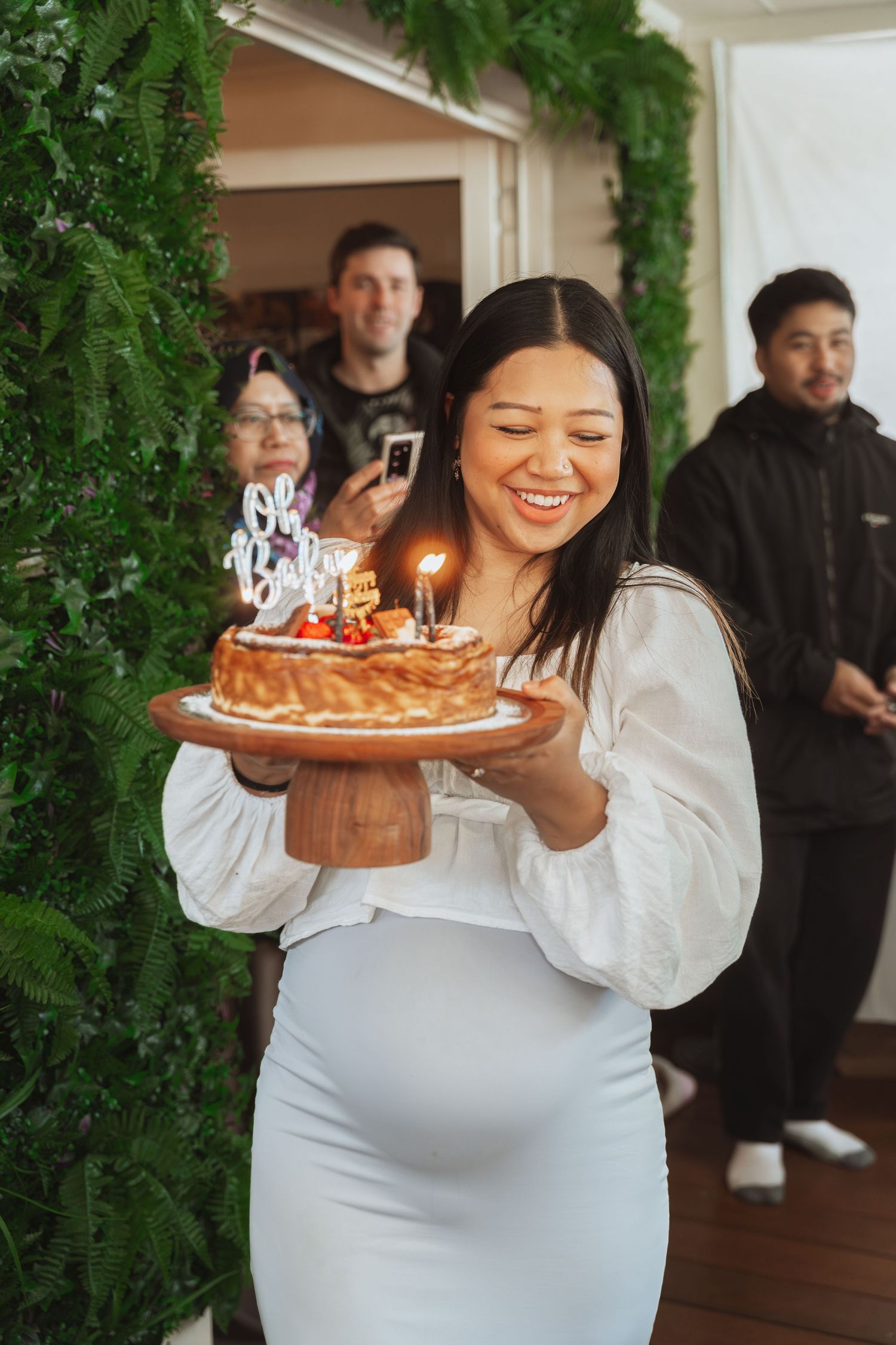 Person in white dress holding birthday cakes with lit candles at an indoor celebration.