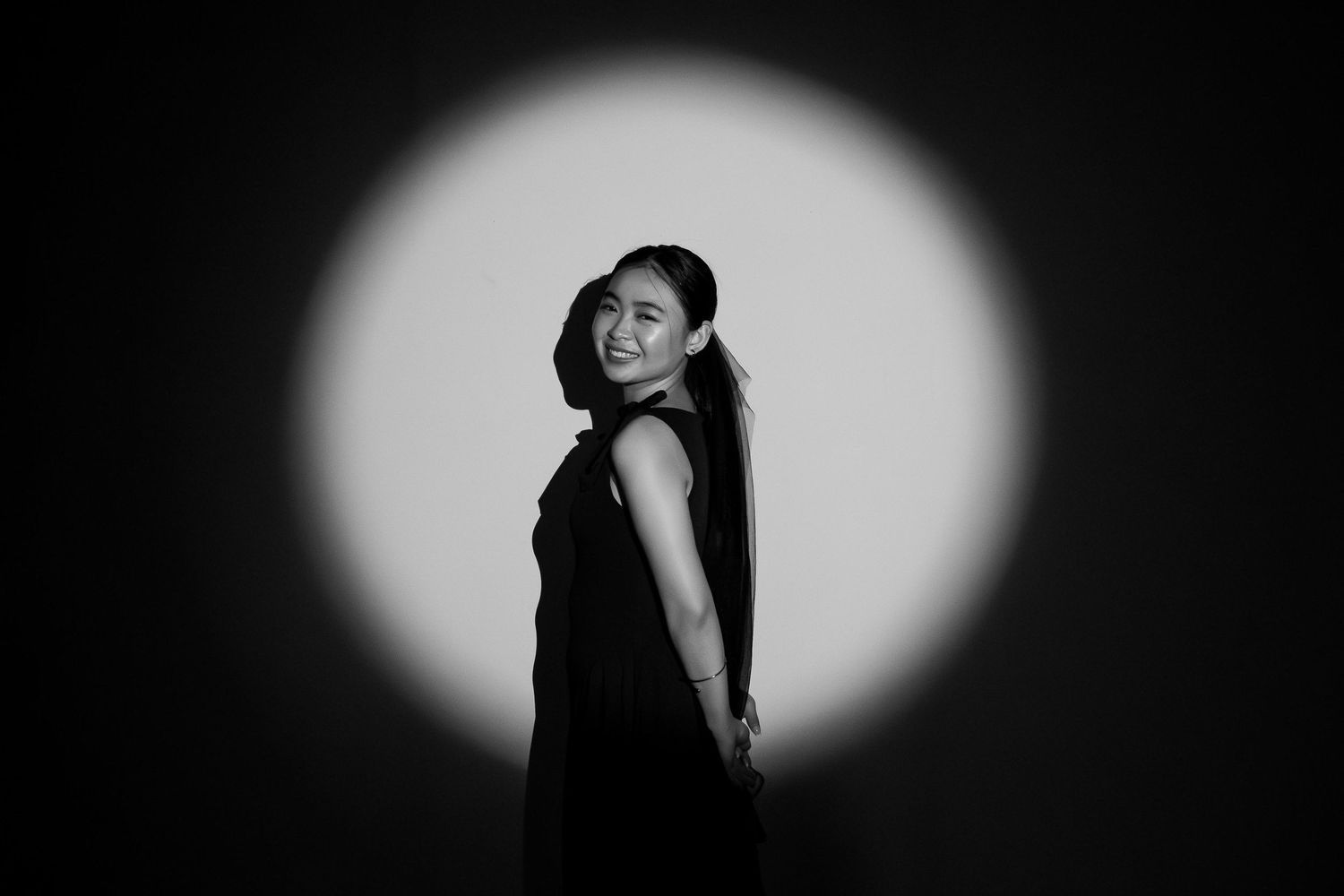 Smiling young woman in black dress posing elegantly in a black and white spotlight.