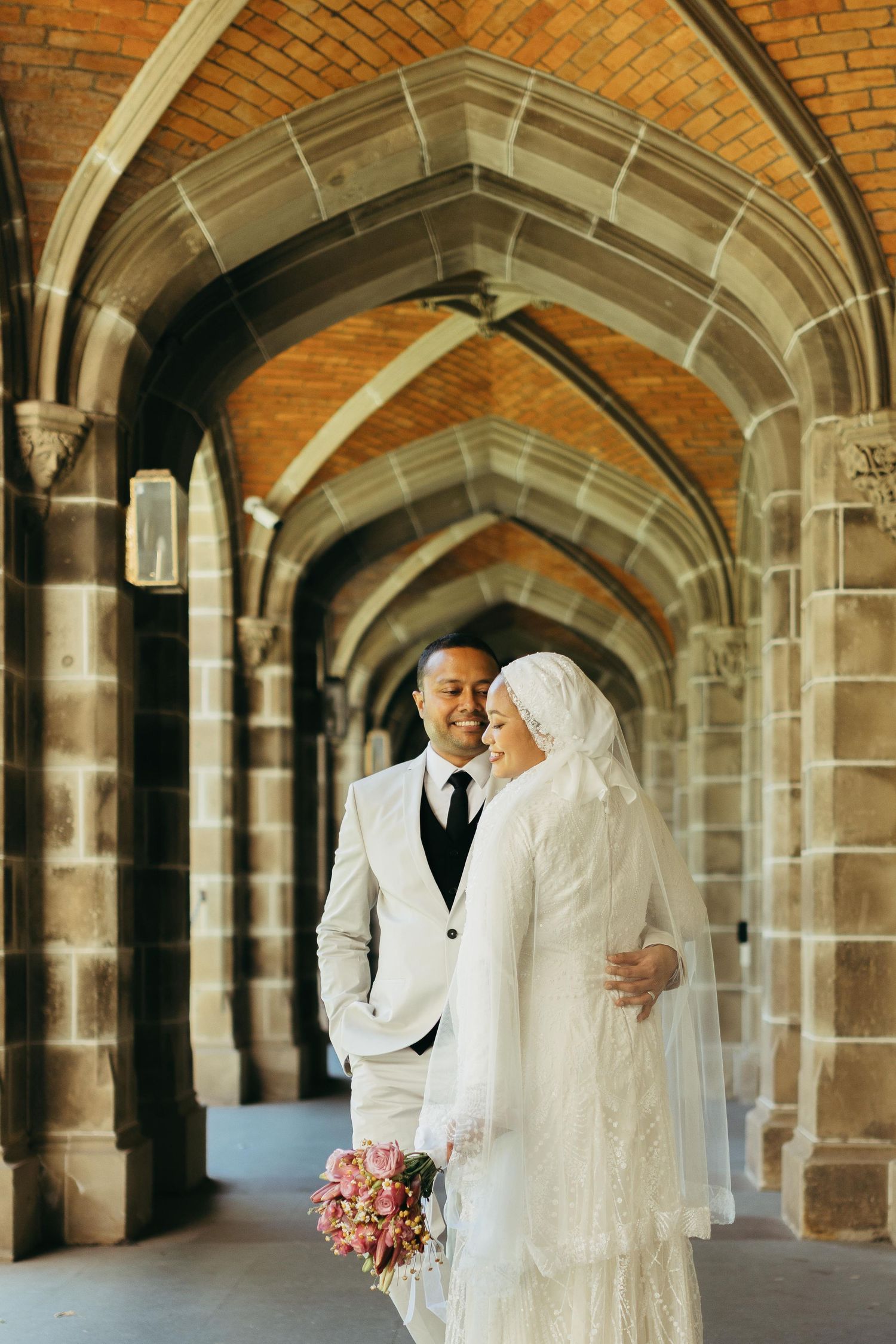 Muslim bride and groom walking under Gothic stone arches, bride wearing white hijab and lace gown with bouquet.