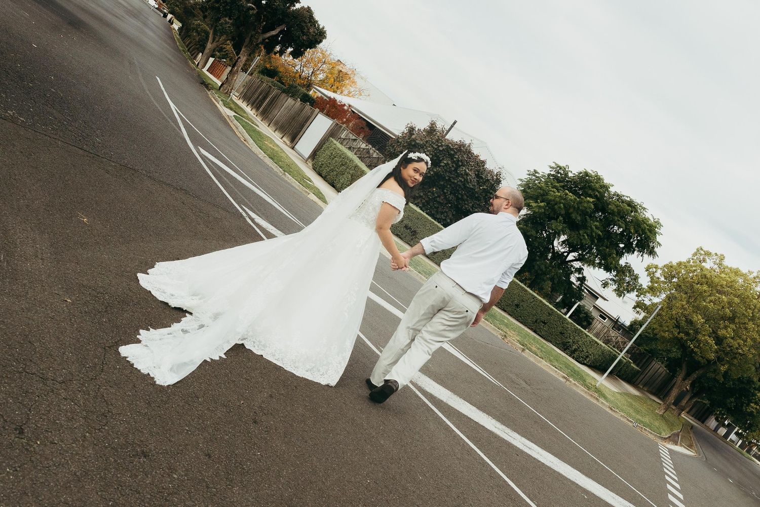 Bride and groom holding hands walking on a road, bride in flowing white gown with long train on their wedding day.