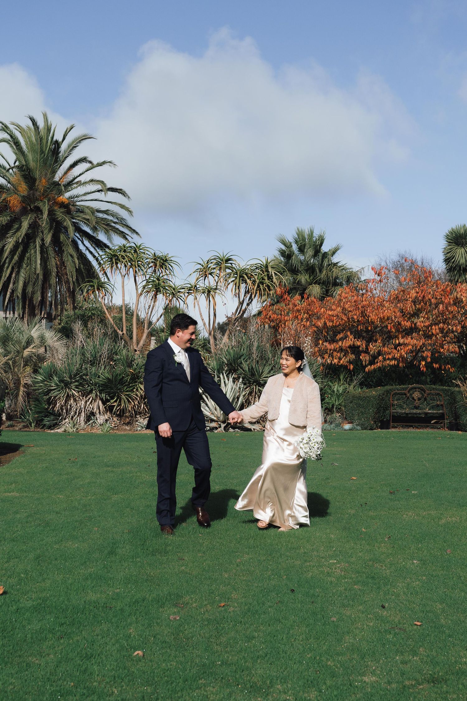 Bride and groom walk hand-in-hand on lush green lawn surrounded by palm trees and colorful gardens on their wedding day.