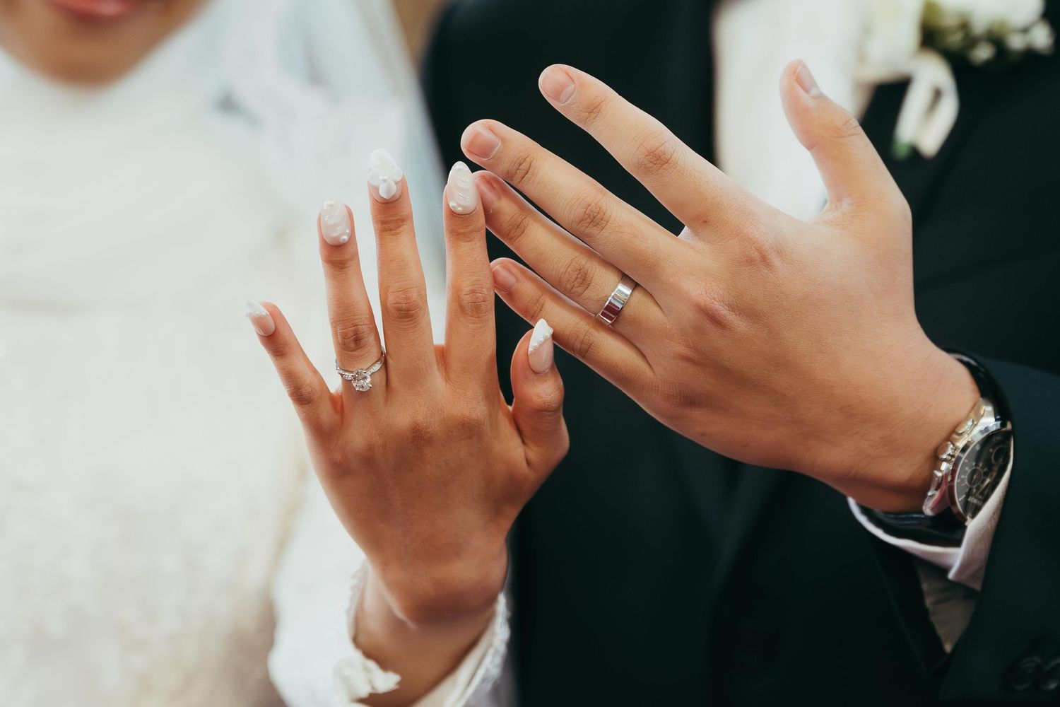 Newlywed couple displaying wedding rings on their fingers, bride in white lace and groom in black suit.
