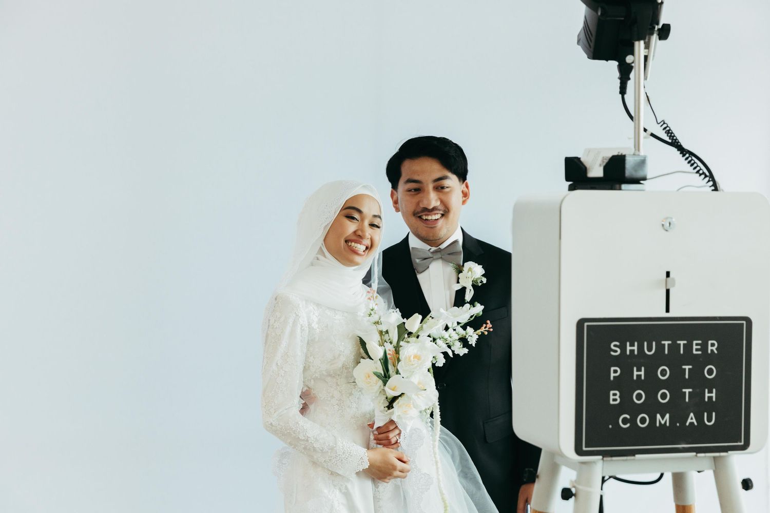 Smiling Muslim bride and groom pose together at a wedding photo booth in white attire.