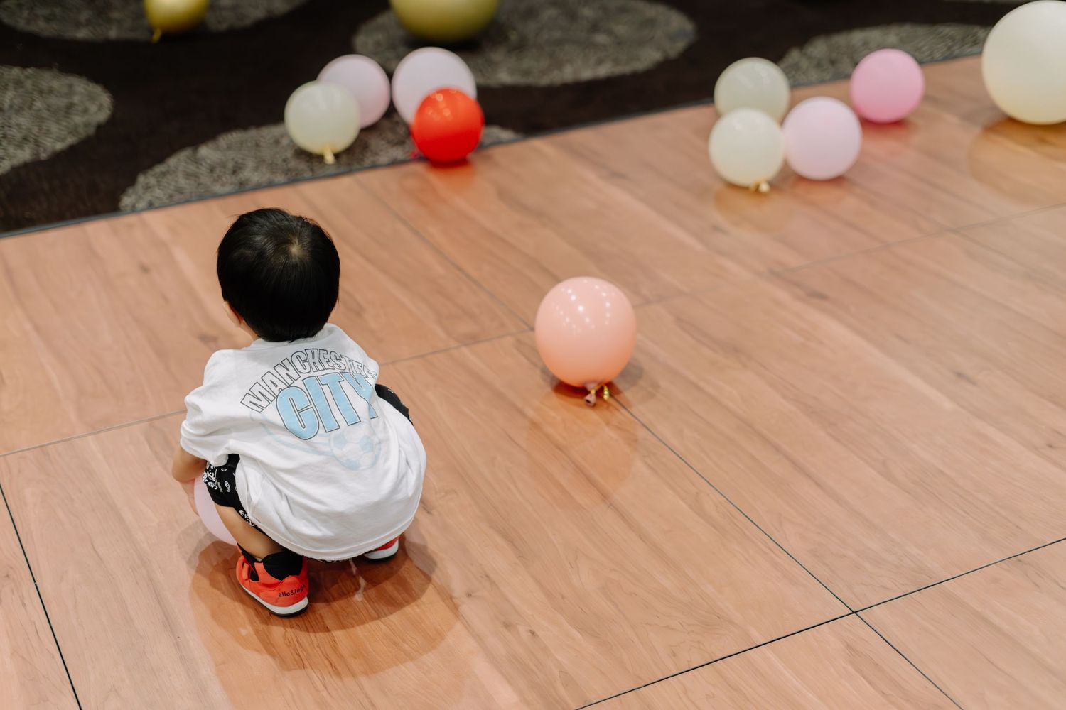 A small child in white sits on a wooden floor with scattered colorful balloons around them.