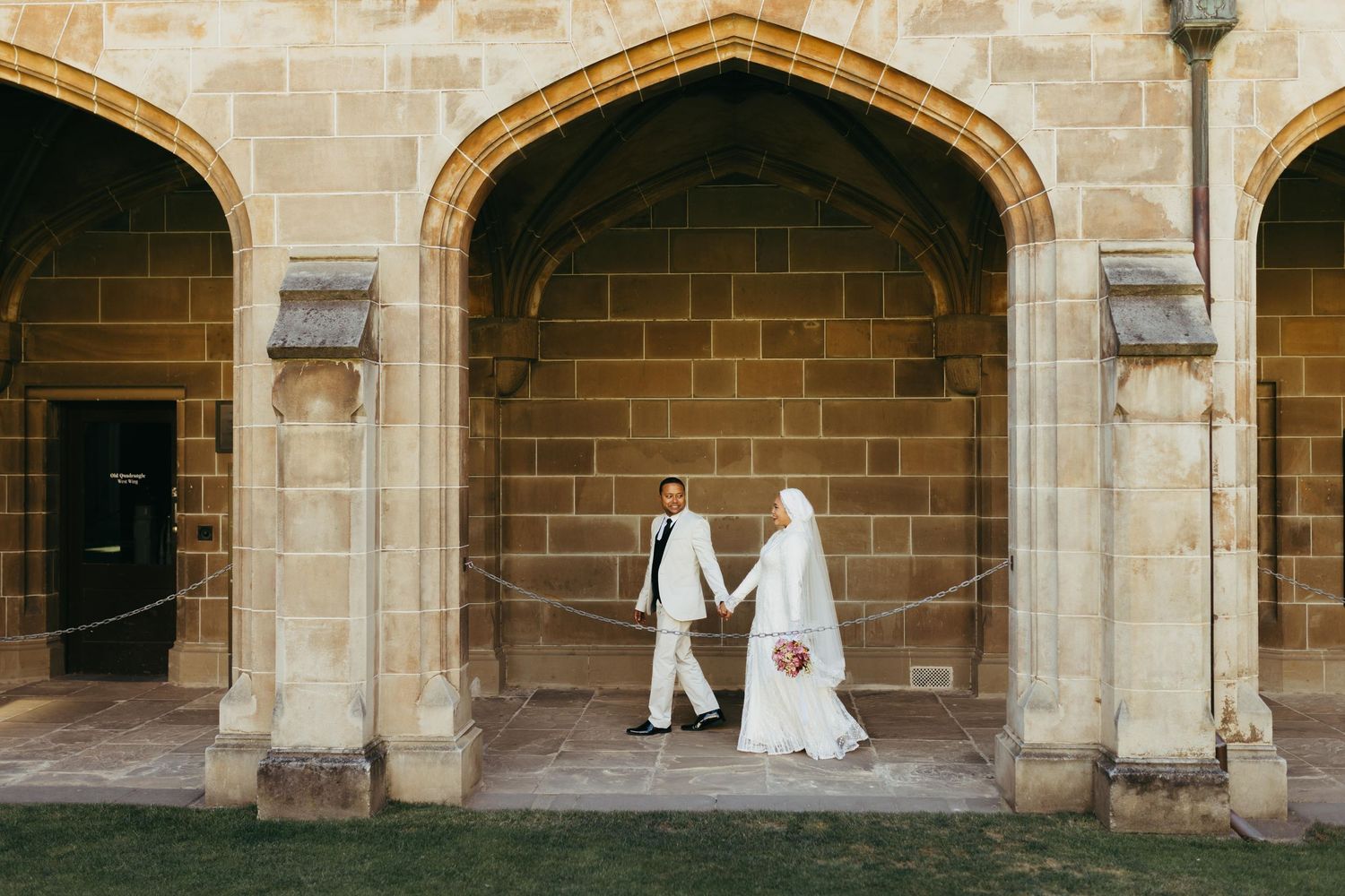 Muslim bride and groom holding hands under stone arches at a historic building on their wedding day.