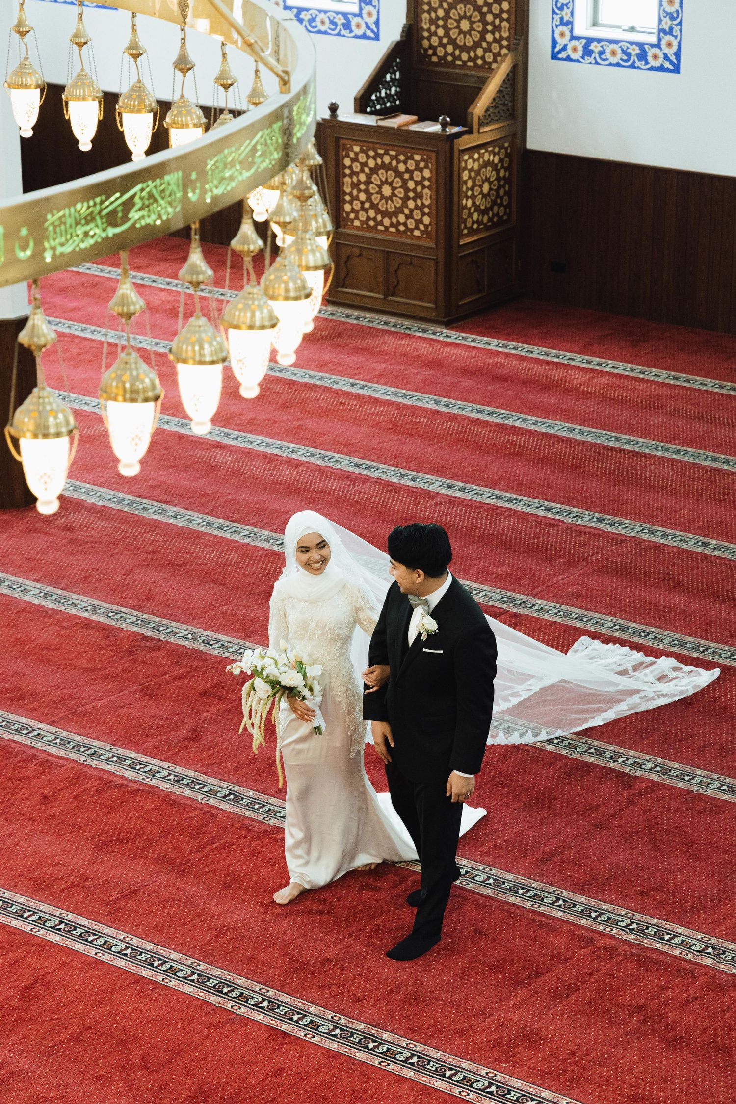 Aerial view of Muslim bride and groom walking on red carpet in ornate mosque during Islamic wedding ceremony.
