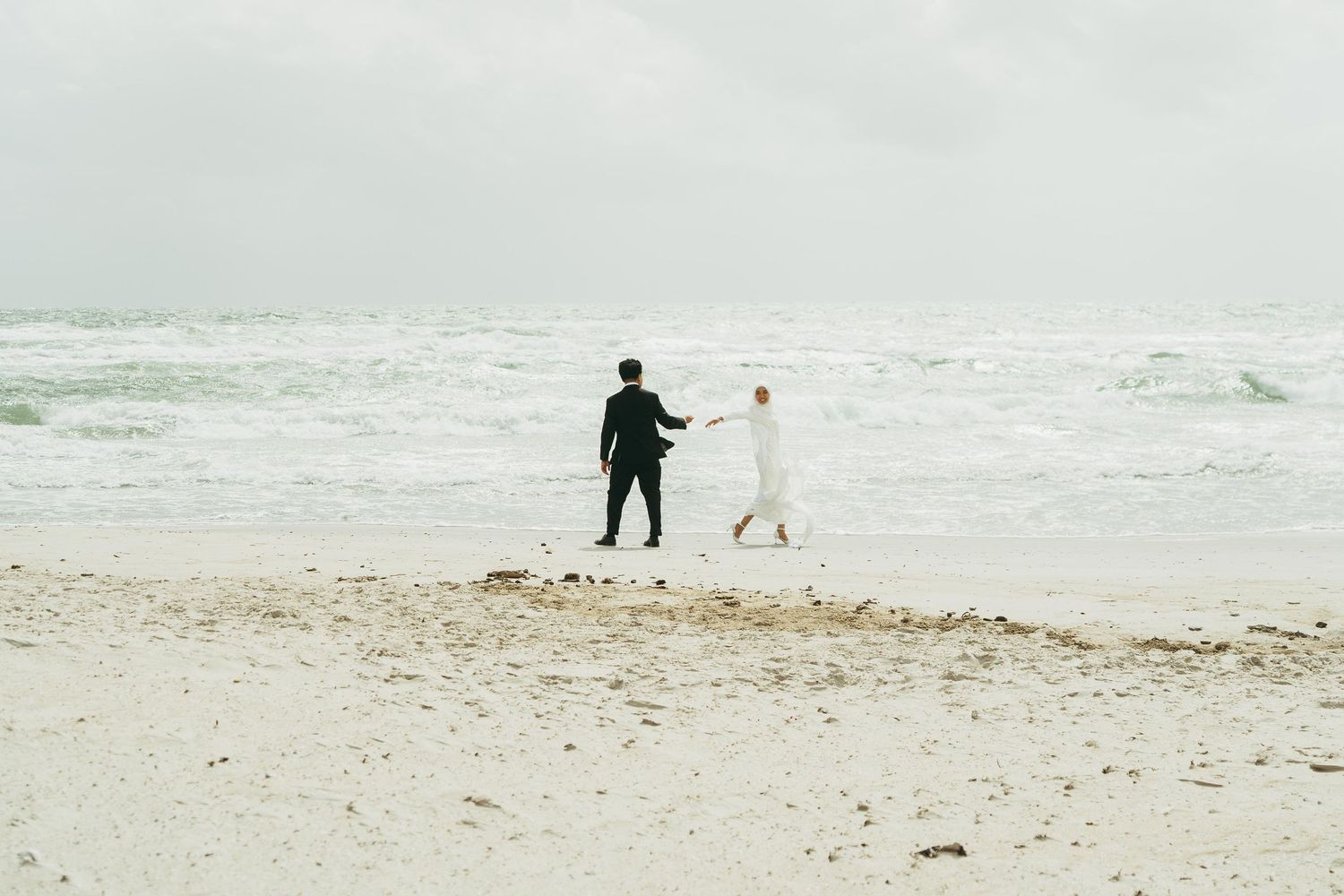 Muslim bride and groom holding hands walking on a beach on overcast day.