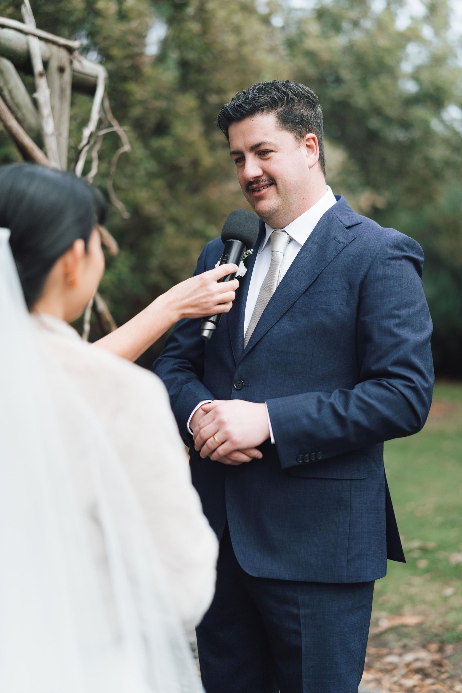 Groom in navy suit smiling while reciting wedding vows outdoors, with bride in white veil holding microphone.