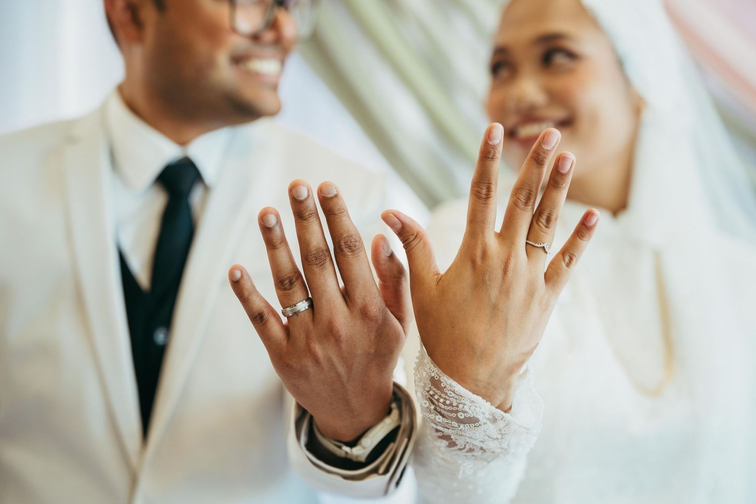 Newlywed couple showing off wedding rings on their hands, smiling in elegant white wedding attire.