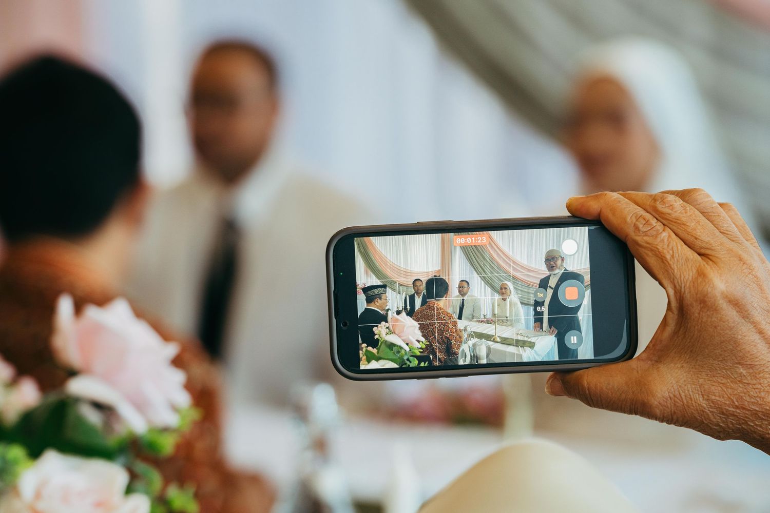 Guest captures wedding ceremony moment on smartphone with floral decorations in foreground.