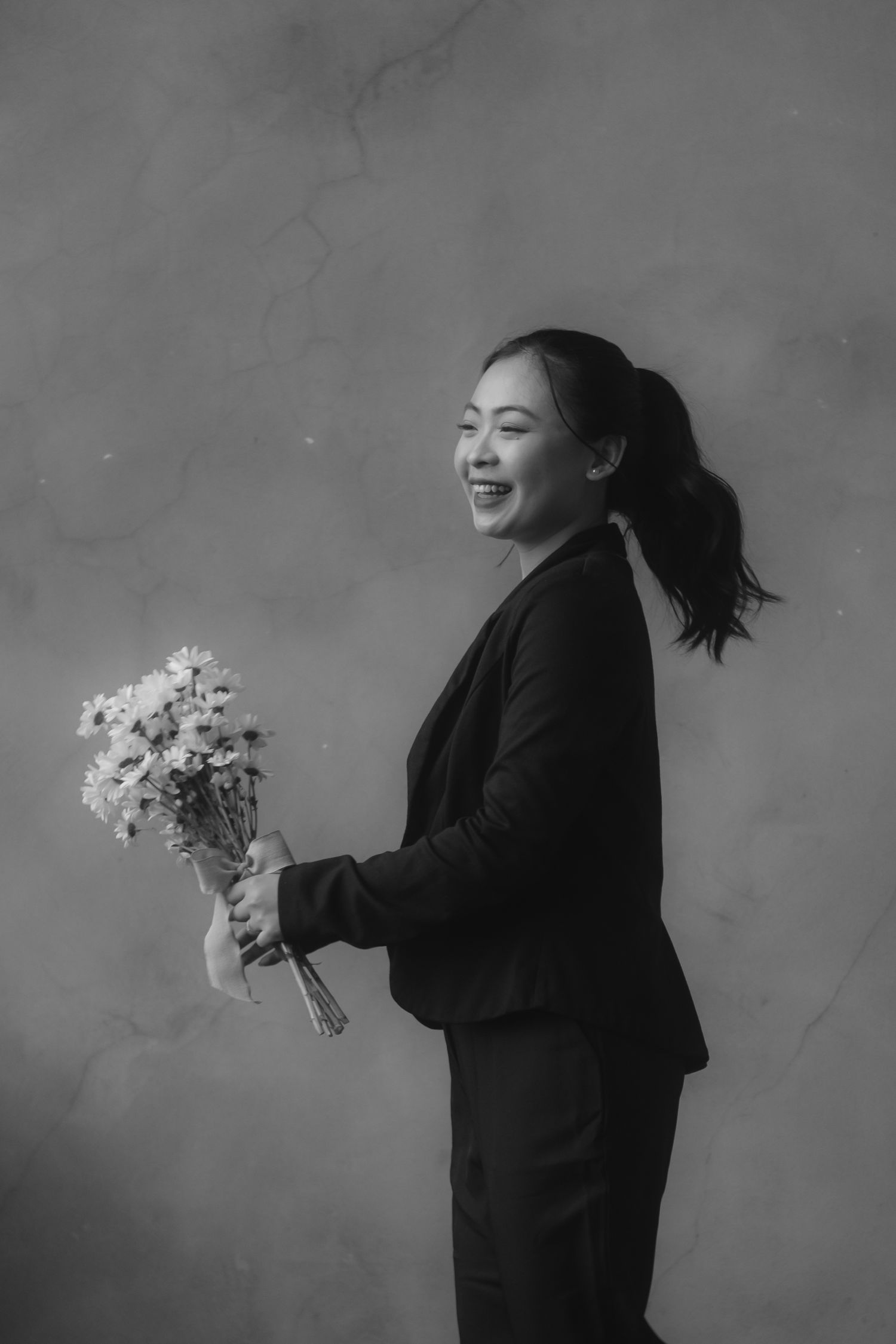 Black and white portrait of a smiling Asian woman in a suit holding a bouquet of flowers against a marble background.