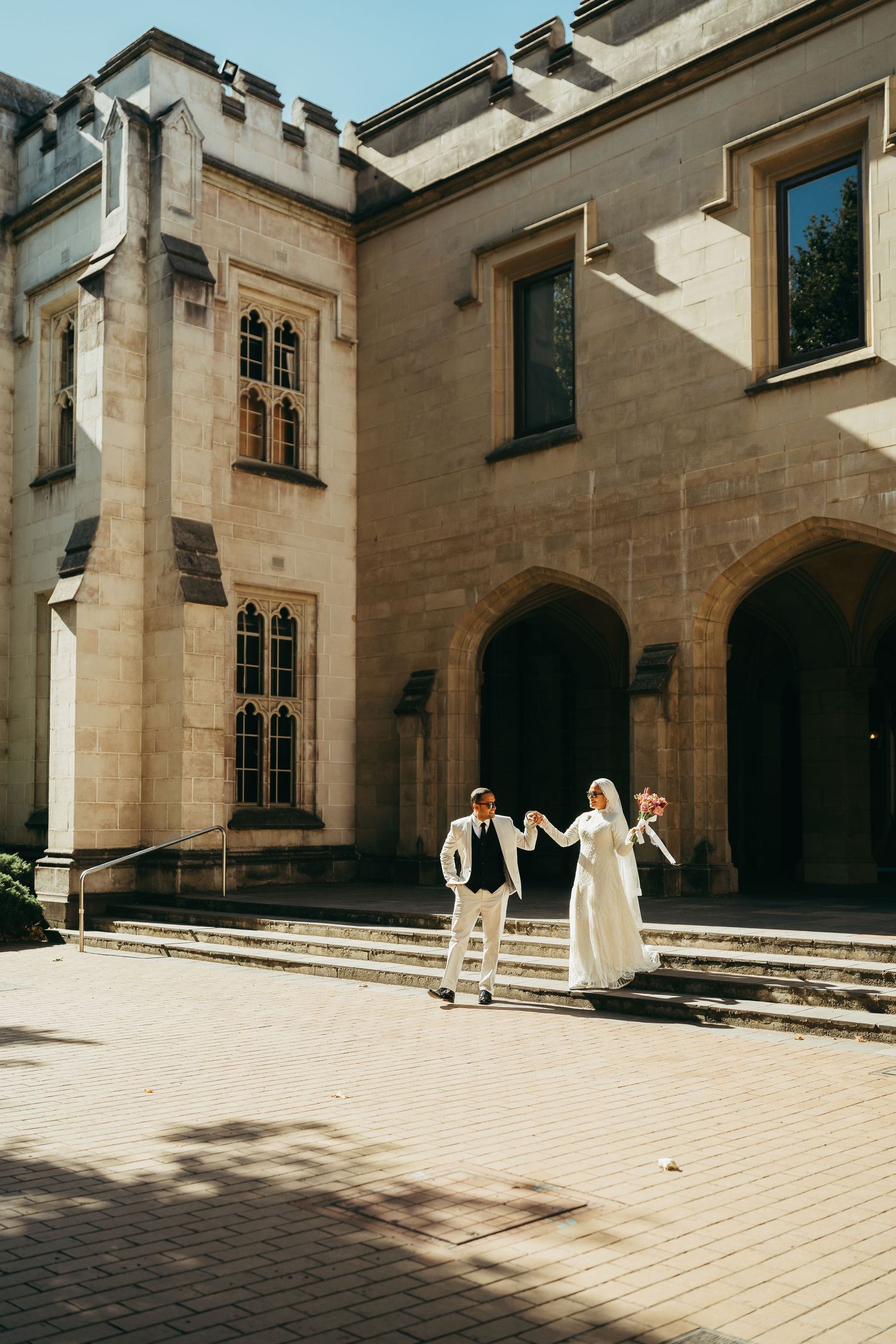 A couple in wedding attire stands on stone steps outside a Gothic-style building with arched doorways and tall windows.