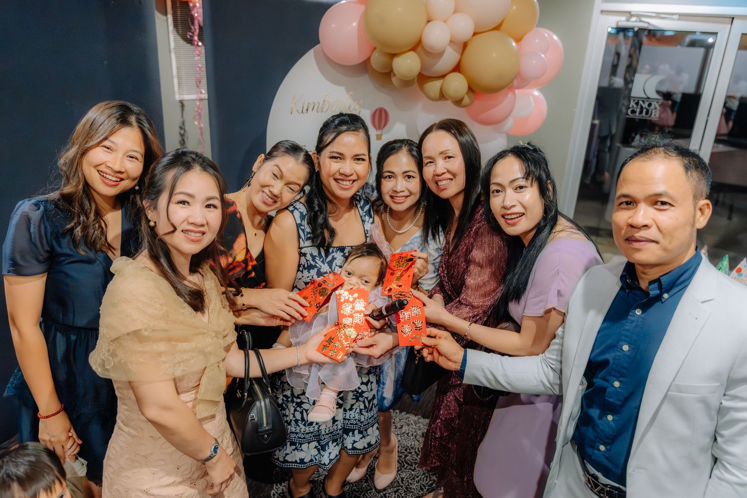 People smile and embrace at a social gathering with pink and beige balloon decorations in the background.