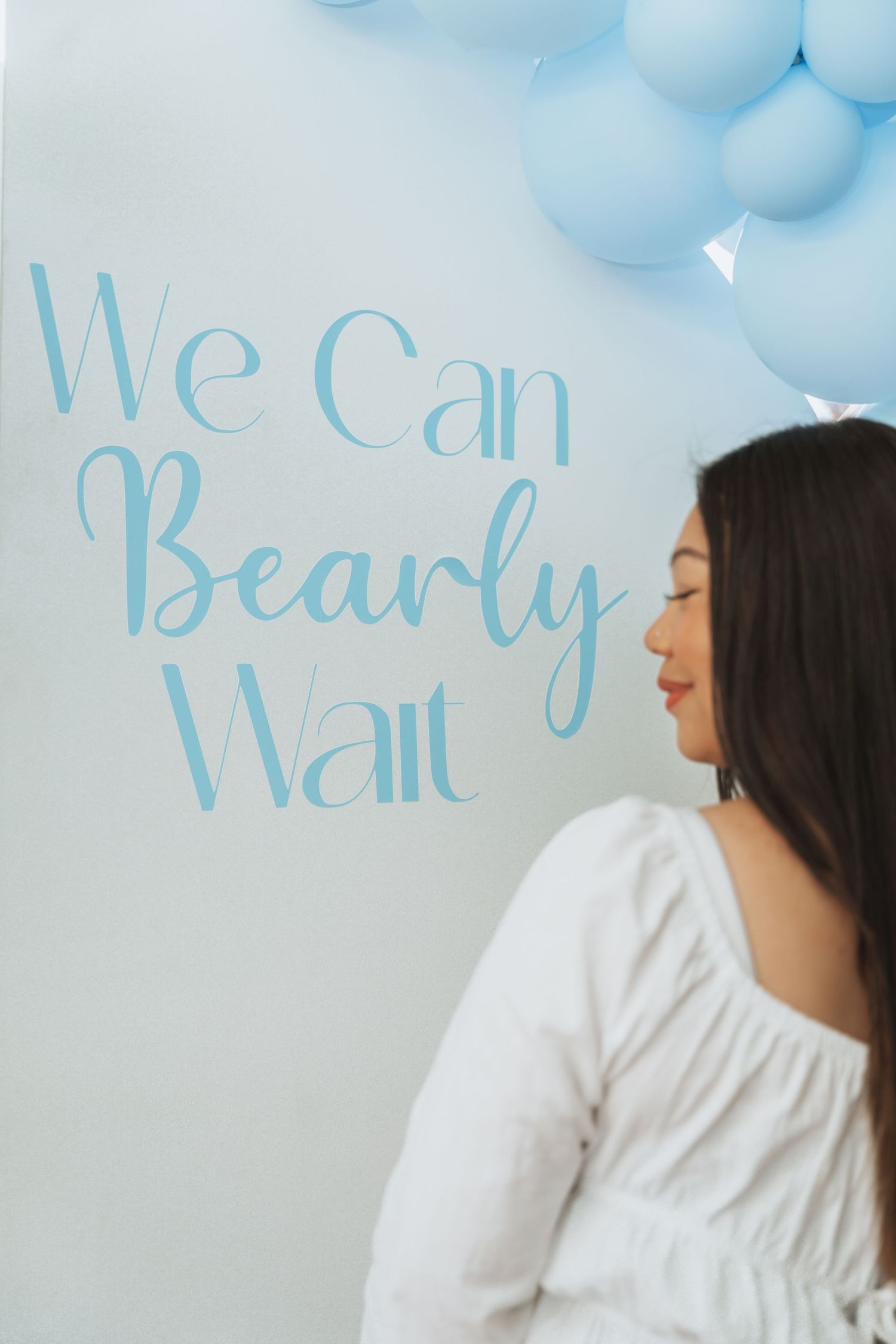 Baby shower decoration with light blue balloons and 'We Can Bearly Wait' text against white wall.