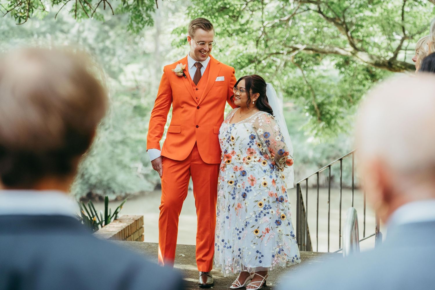 Groom in bold orange suit smiles with bride in floral dress during outdoor wedding ceremony.