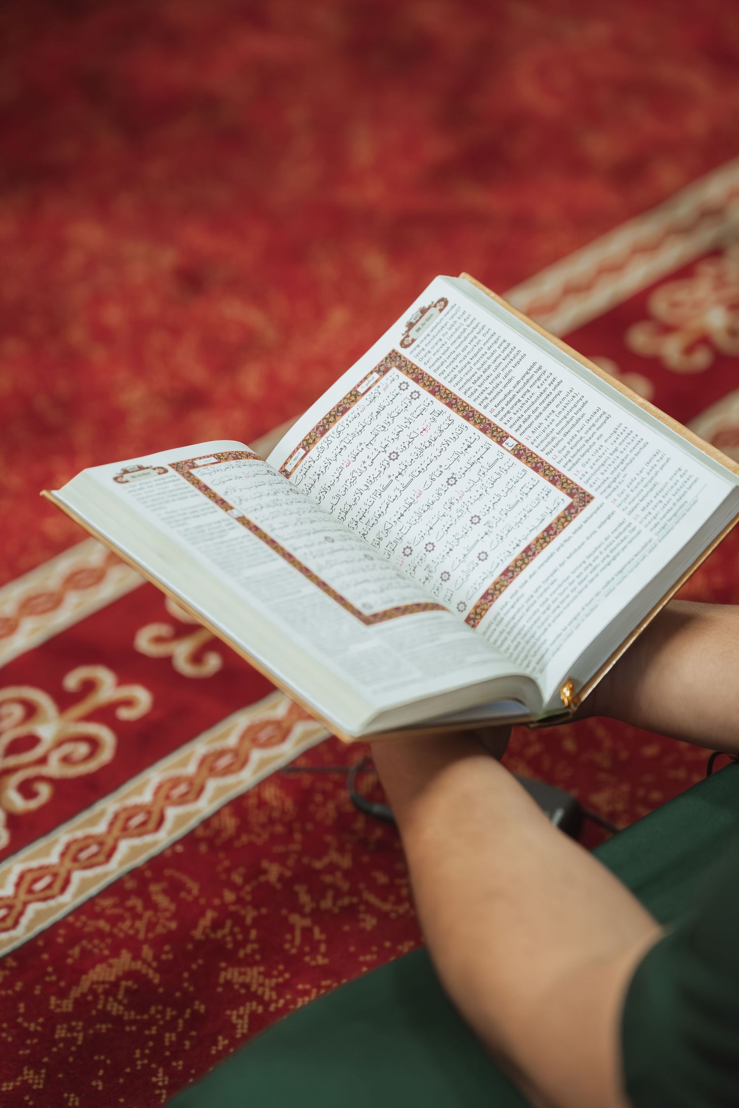 Person sitting on a red patterned prayer rug reading the Quran in a mosque.