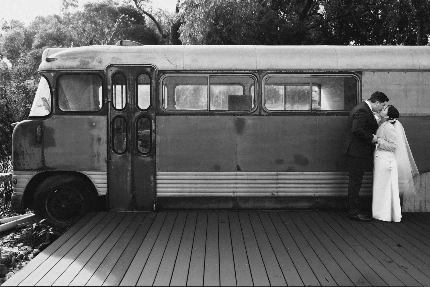 Bride and groom share a romantic embrace beside a vintage bus in a black and white wedding photo.