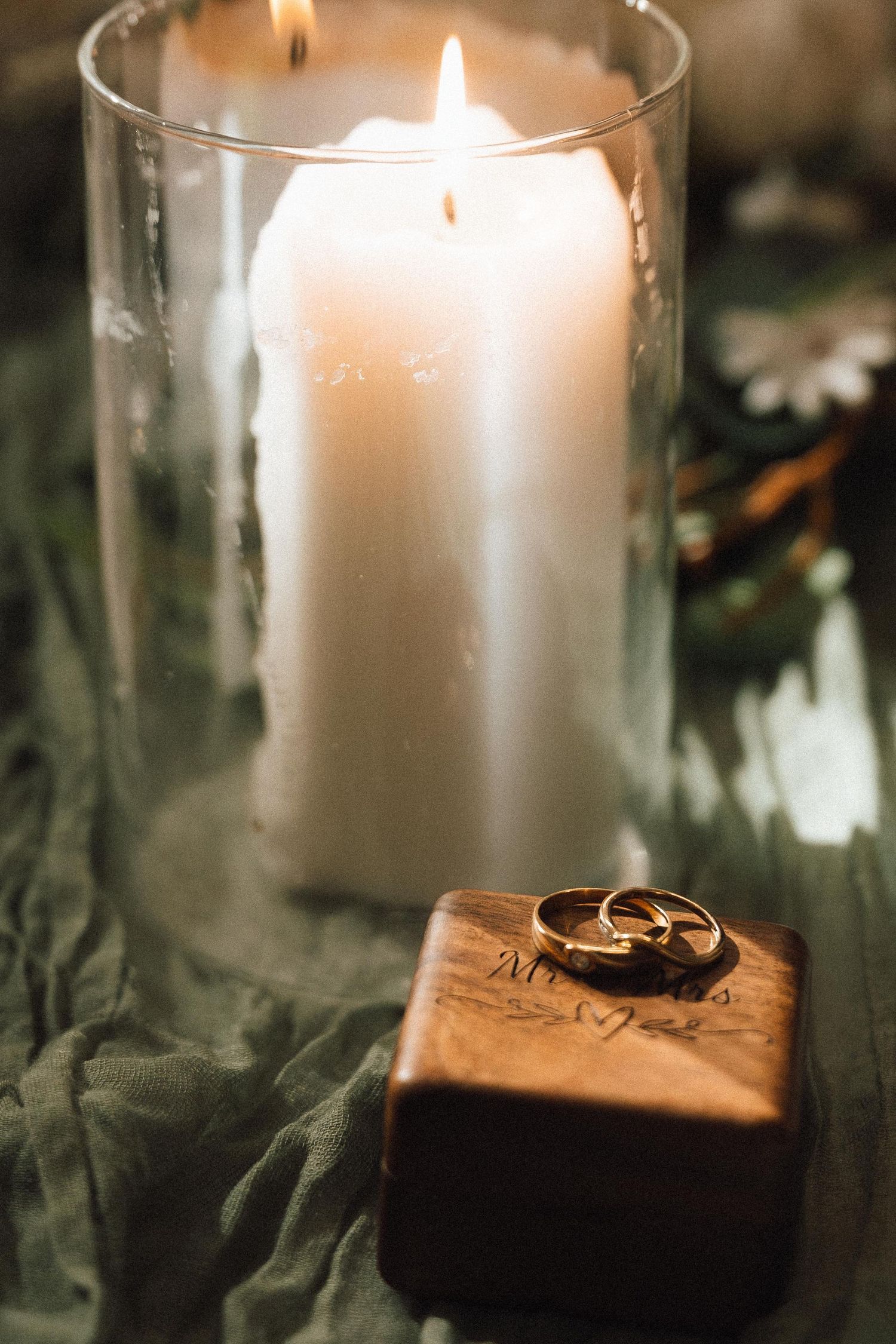 Gold wedding rings resting on a wooden box beside a glowing white pillar candle in a glass holder.