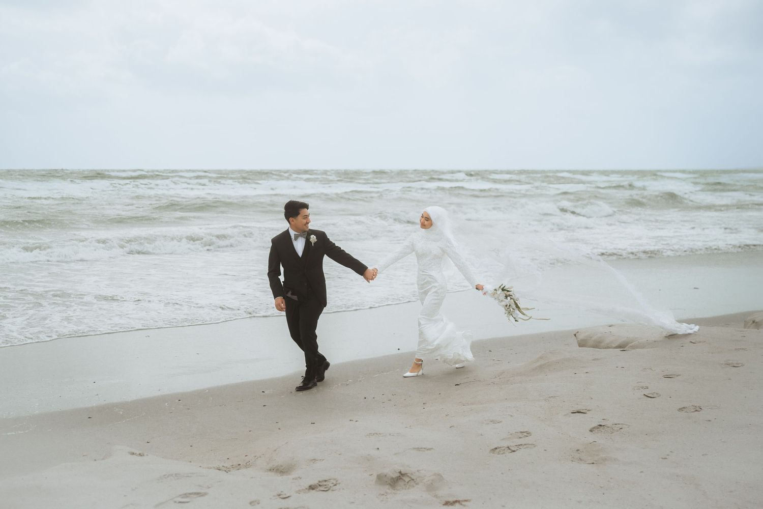 Wedding couple walking along a misty beach shoreline in formal attire.