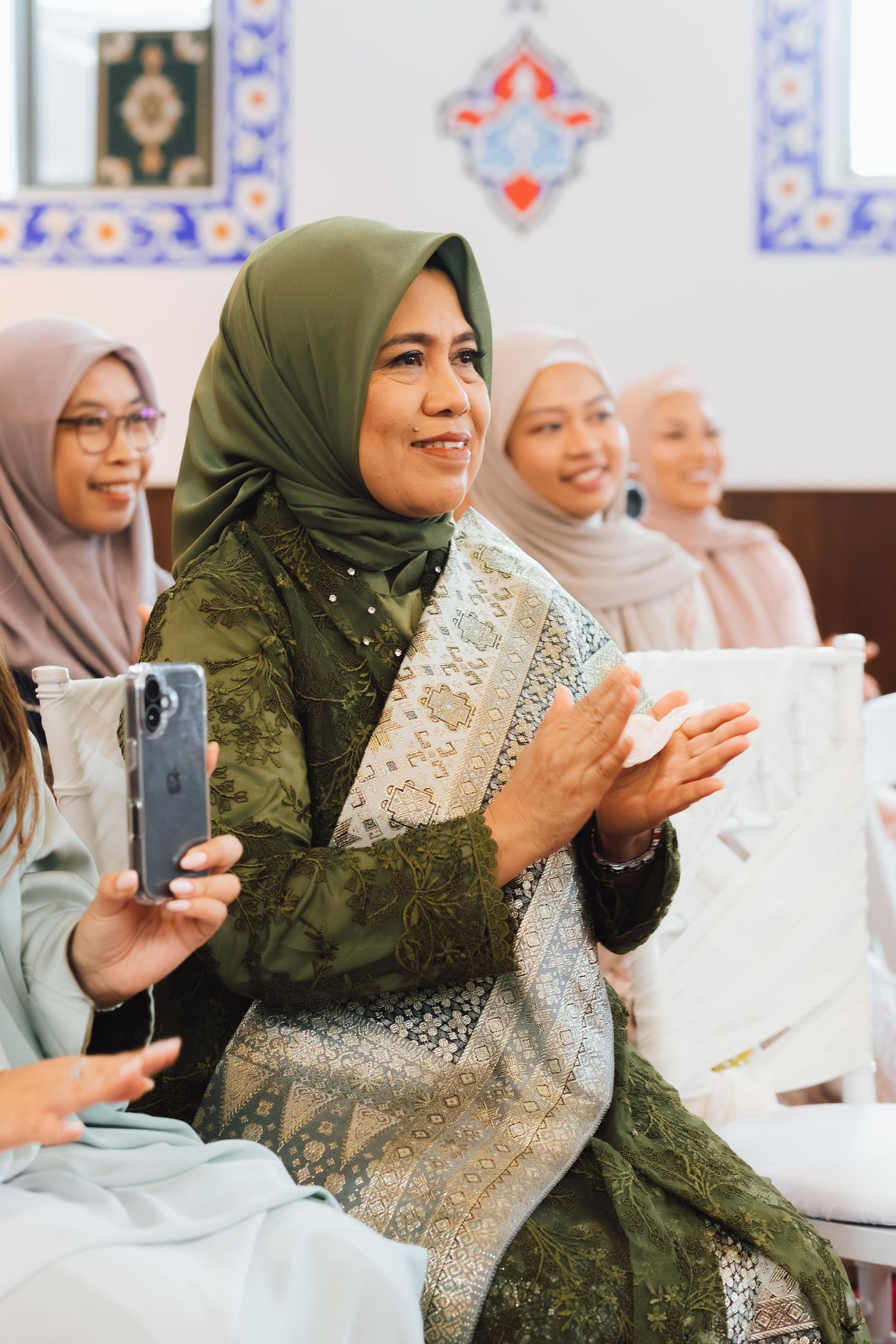 Smiling Muslim women in hijabs clapping at a wedding ceremony indoor.