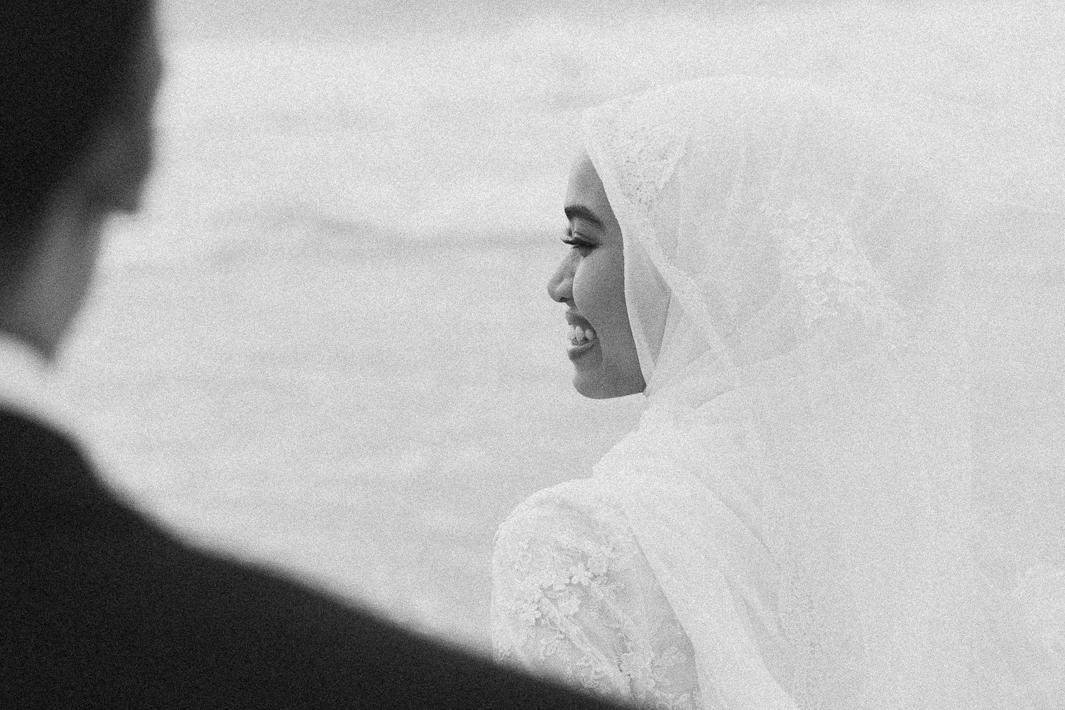 Black and white portrait of smiling Muslim bride in white lace hijab and veil at beach wedding.
