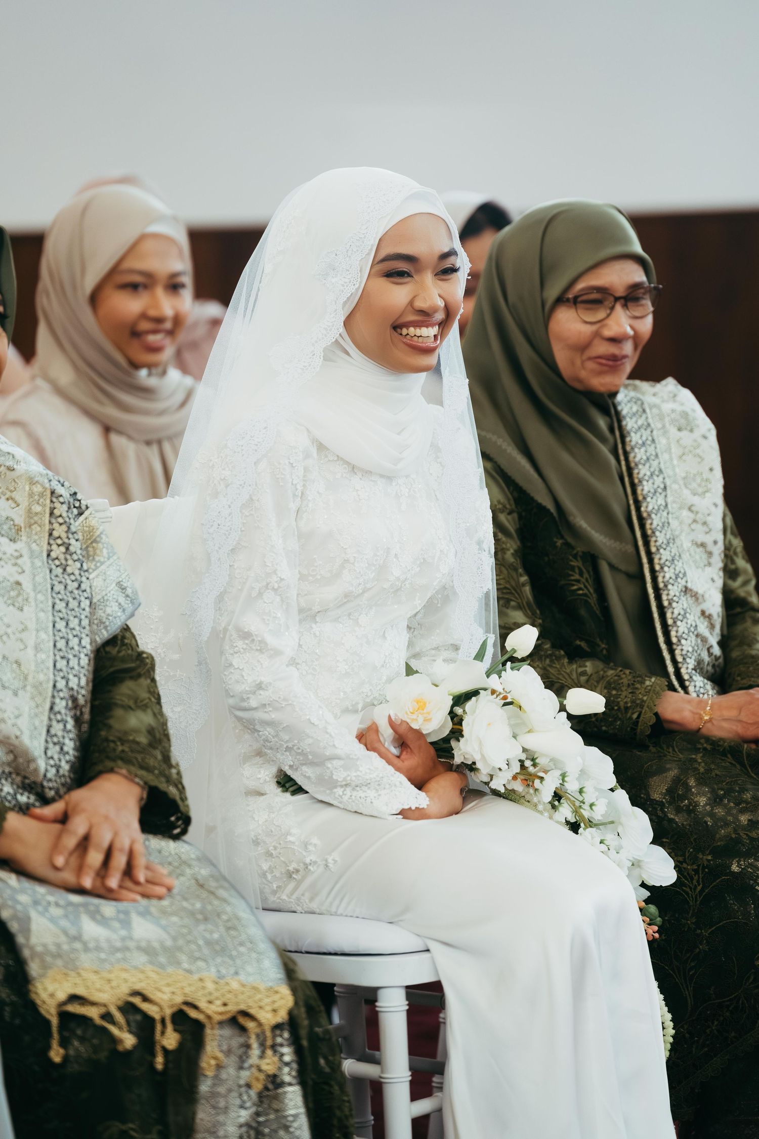 Smiling Muslim bride in white lace hijab gown holding white bouquet seated with family.