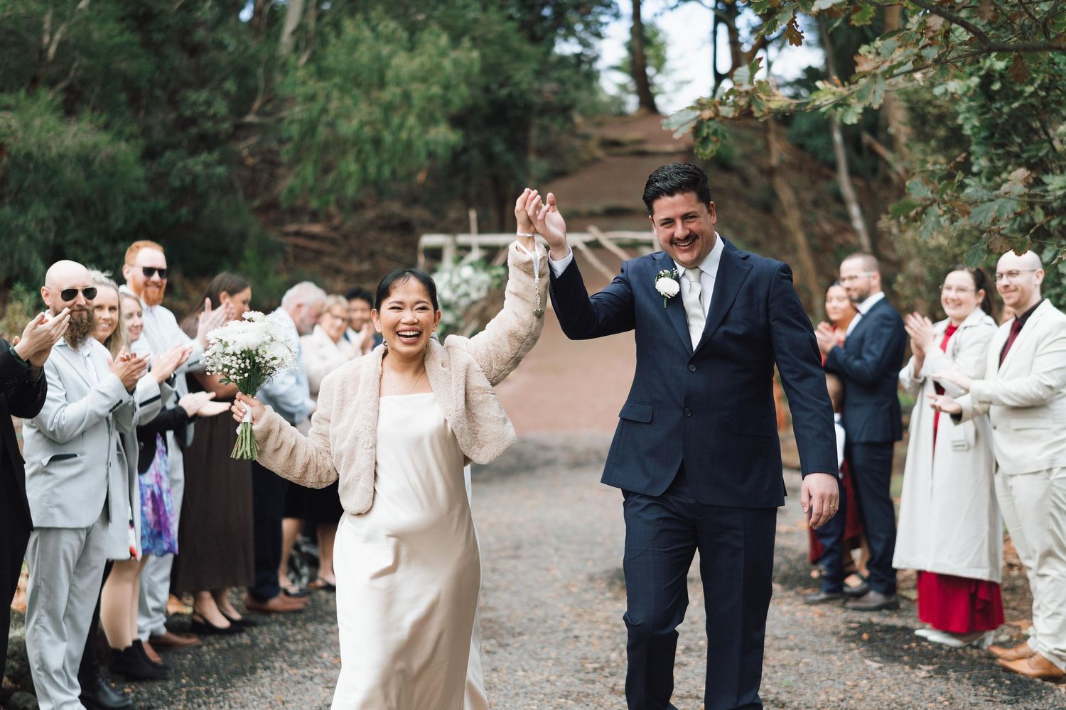 Joyful newlyweds raise hands as guests applaud during outdoor wedding recessional.