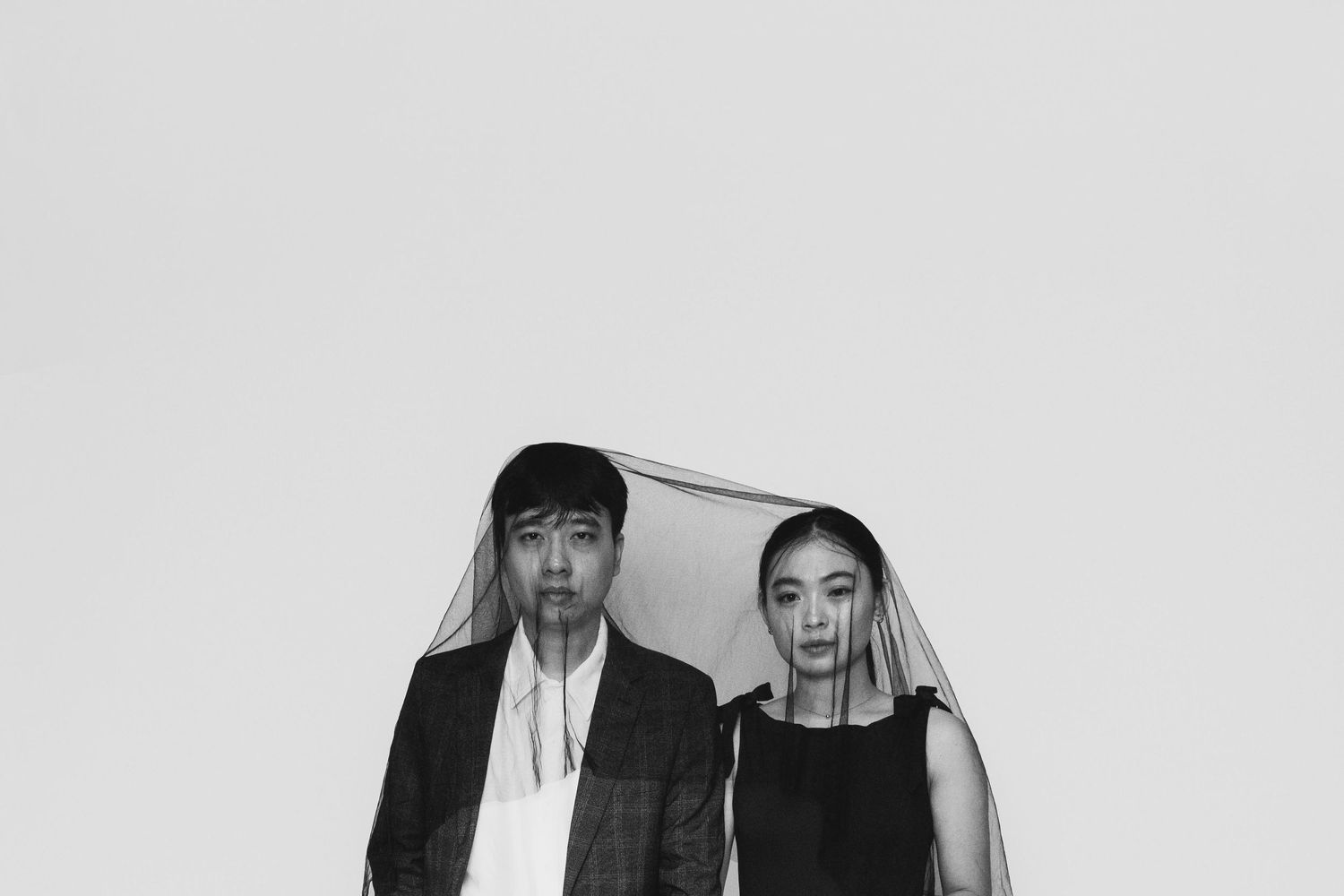 Black and white portrait of a man and woman standing together under a sheer veil against a light background.
