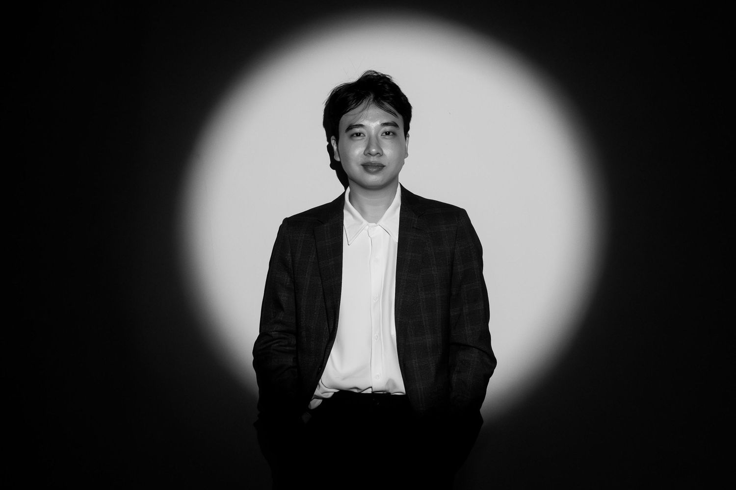 Black and white portrait of a man in a blazer seated against a dramatic spotlight background.