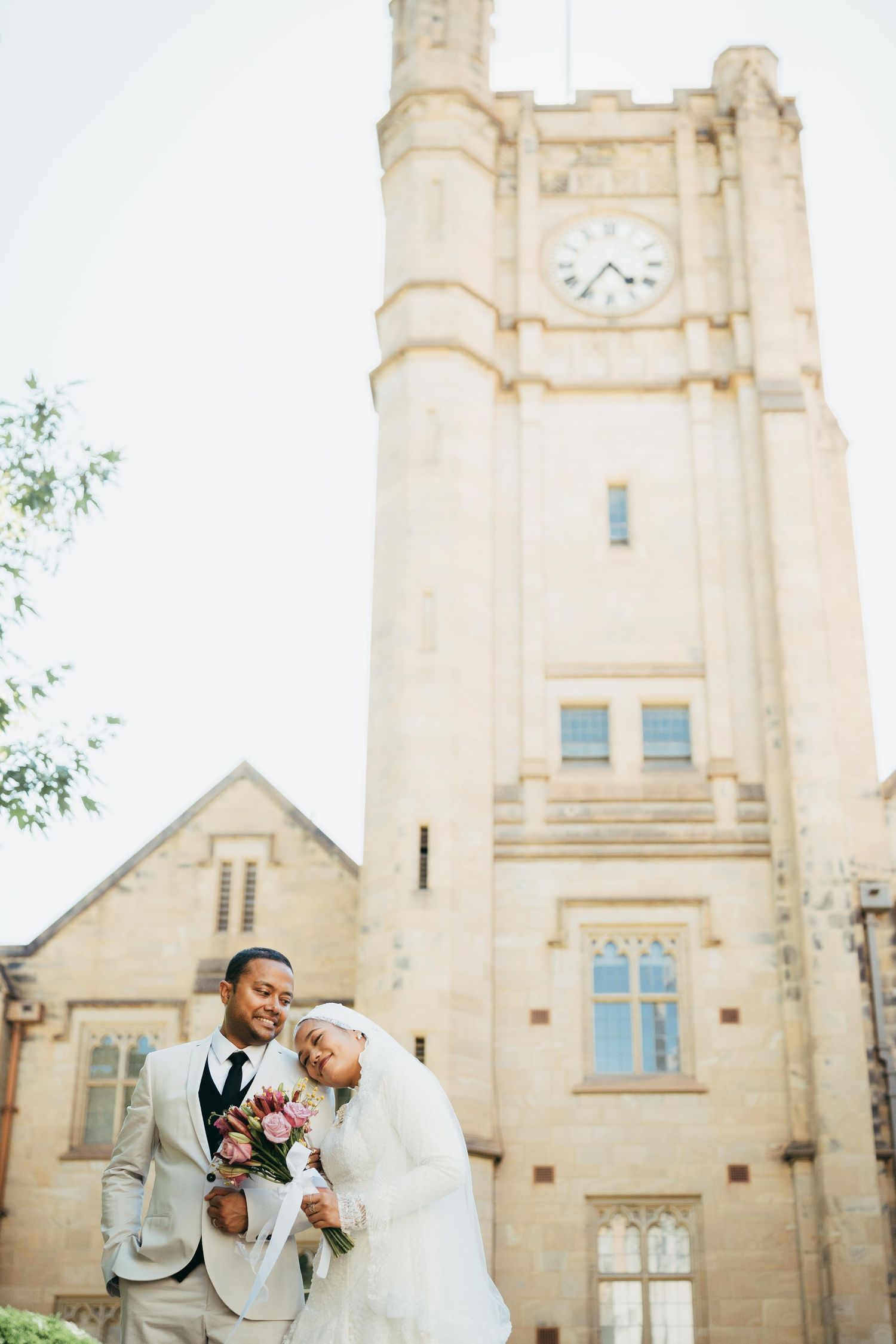 Muslim bride and groom pose romantically before historic stone clock tower with pink bridal bouquet.