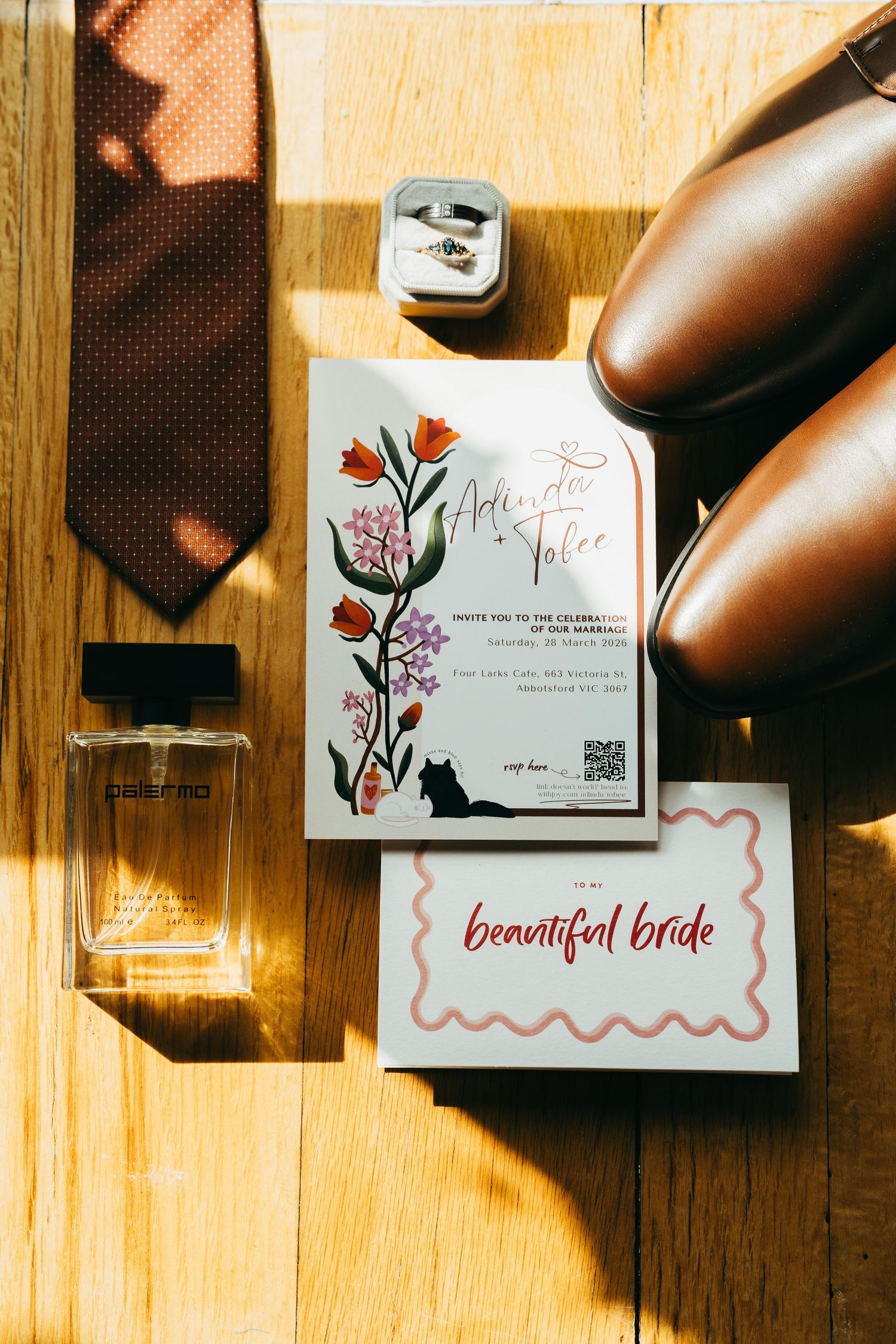 Flat lay of wedding details including floral invitation, cologne, brown leather shoes, tie, and rings on wood.