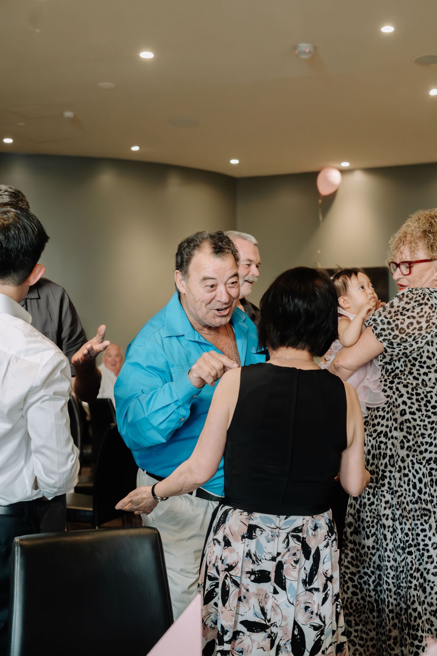 People dancing and socializing at a celebration event with recessed lighting and balloon decorations.