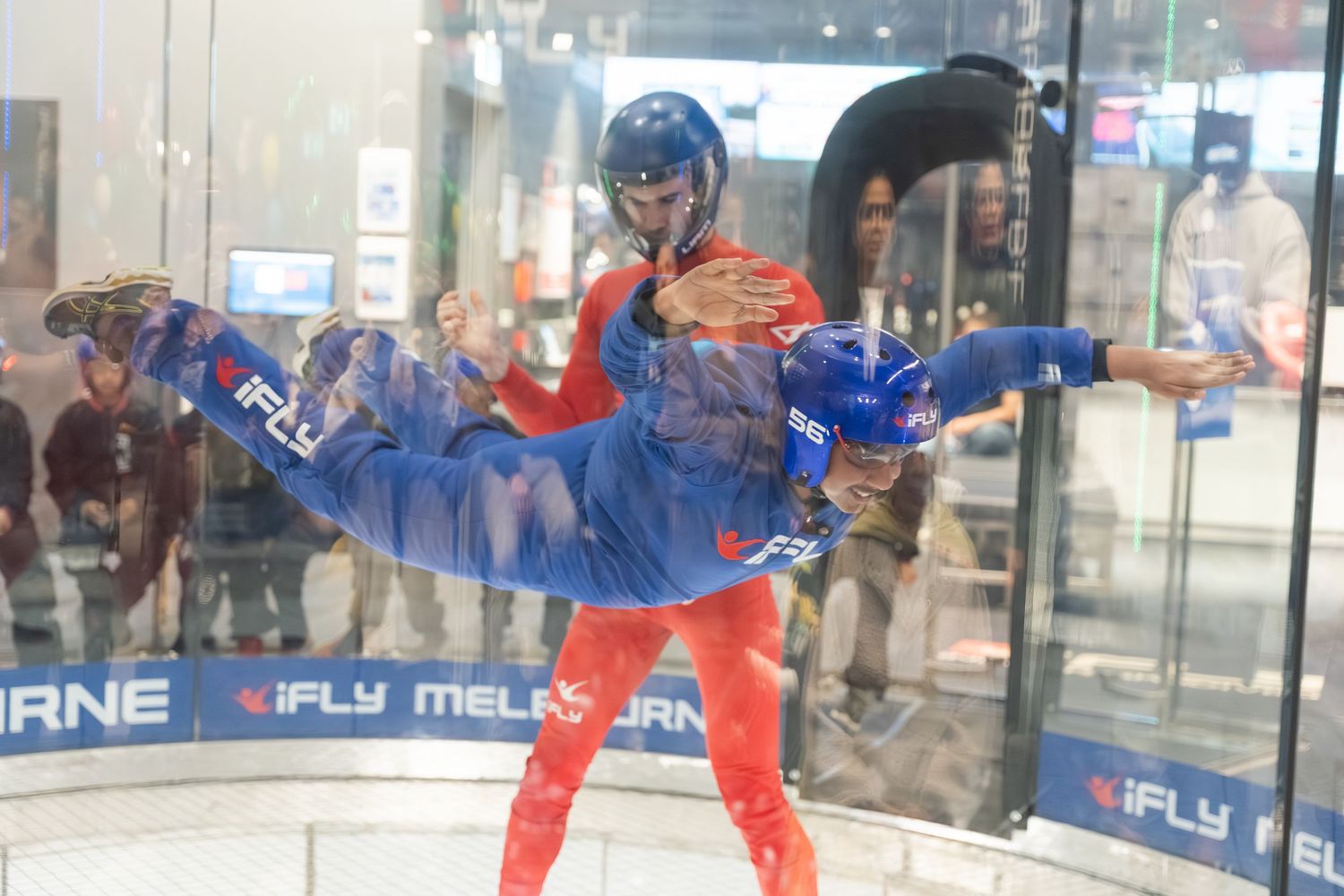 An indoor skydiver performs a horizontal pose in a wind tunnel while wearing a red and blue flight suit.