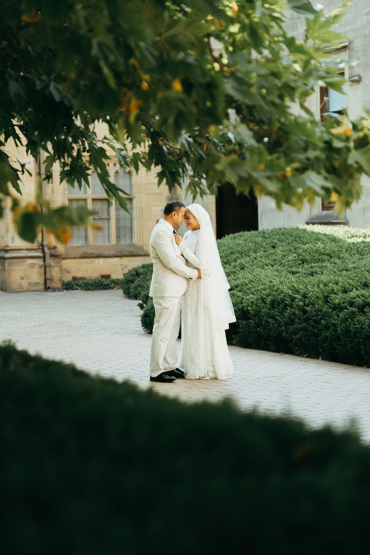 A couple in white wedding attire embraces in a garden setting with lush greenery and a white brick wall backdrop.