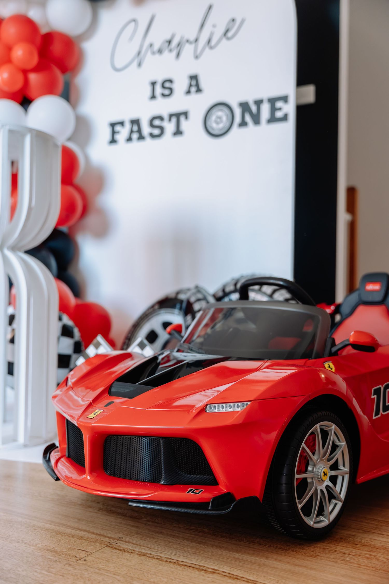 Red toy Ferrari sports car displayed next to white and red balloon decorations and a sign.