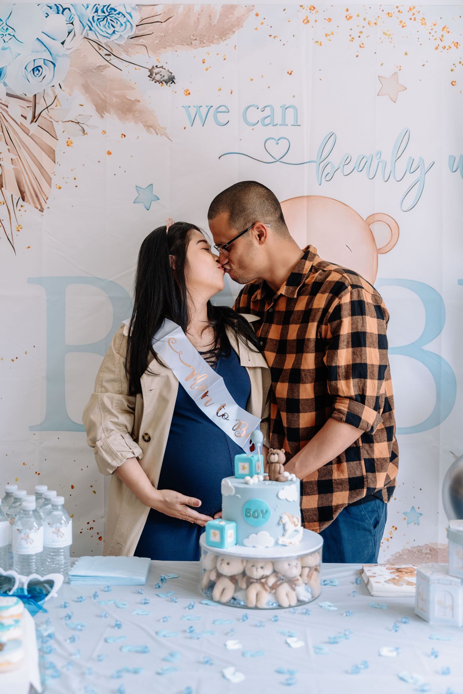 A couple shares an intimate moment during a gender reveal party with a blue and white decorated cake and backdrop.