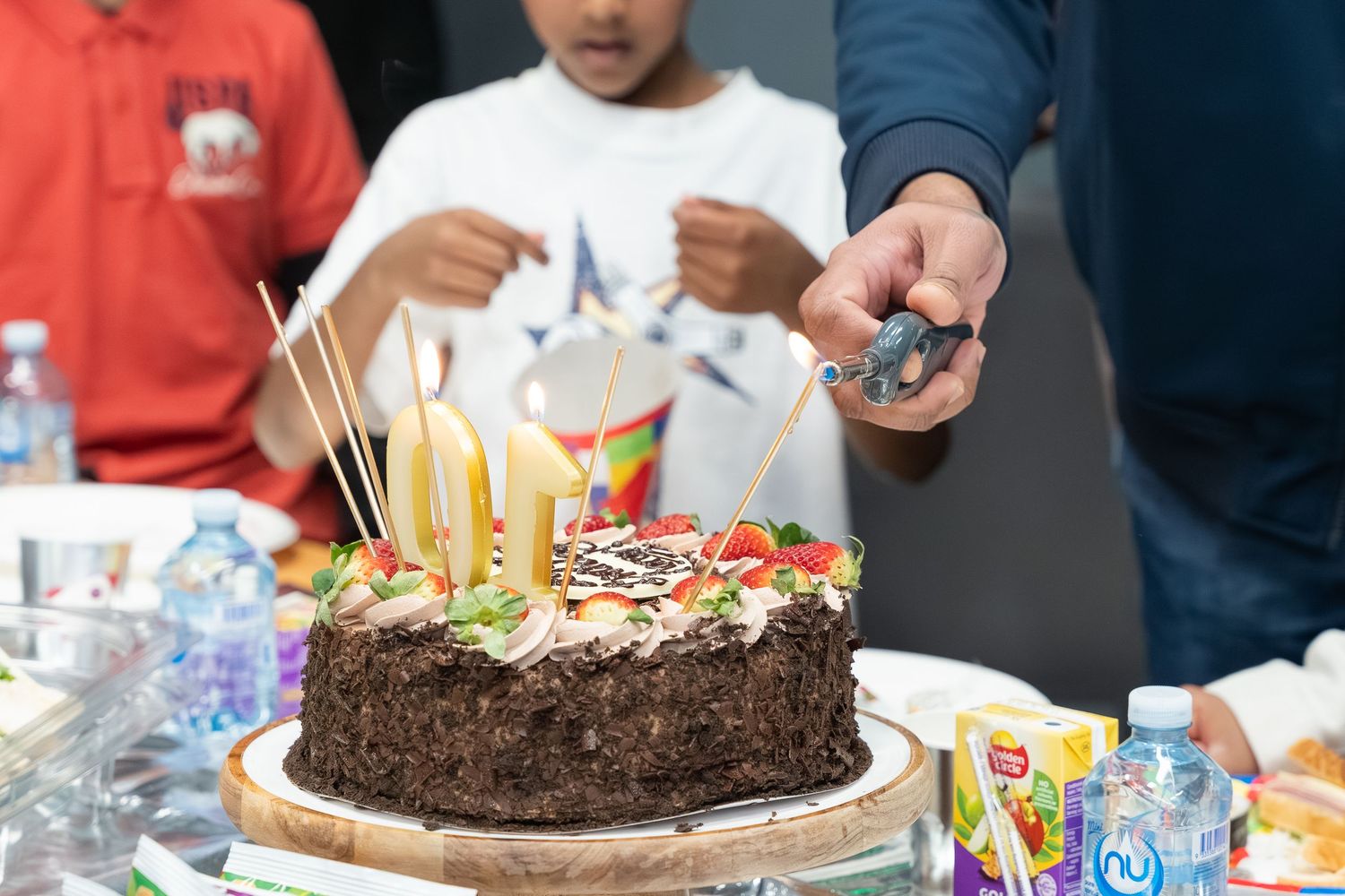 A chocolate birthday cake with lit candles sits on a table surrounded by party decorations and drinks.
